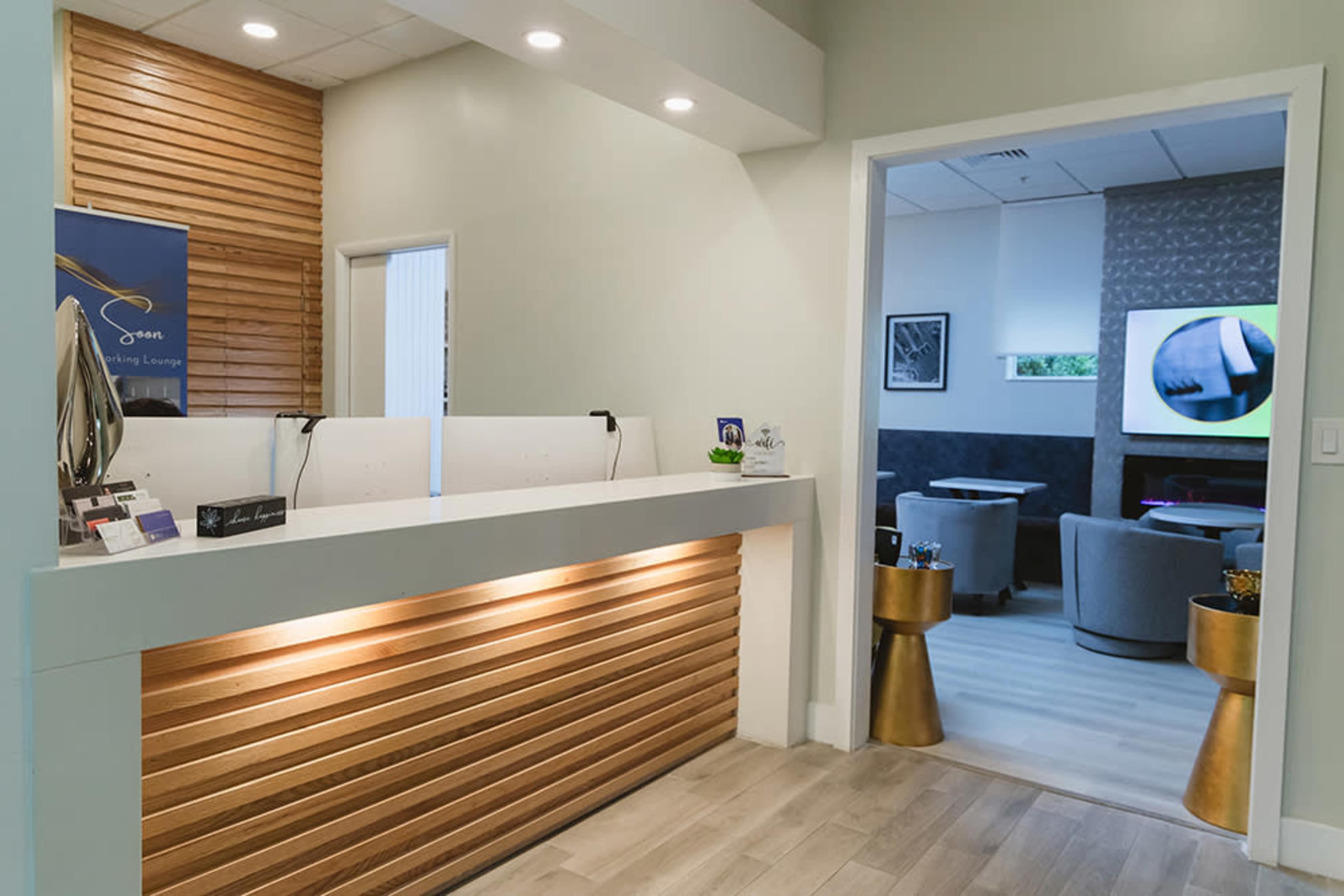 A modern reception area with a wooden front desk and a doorway leading to a lounge space featuring gray seating and a TV.