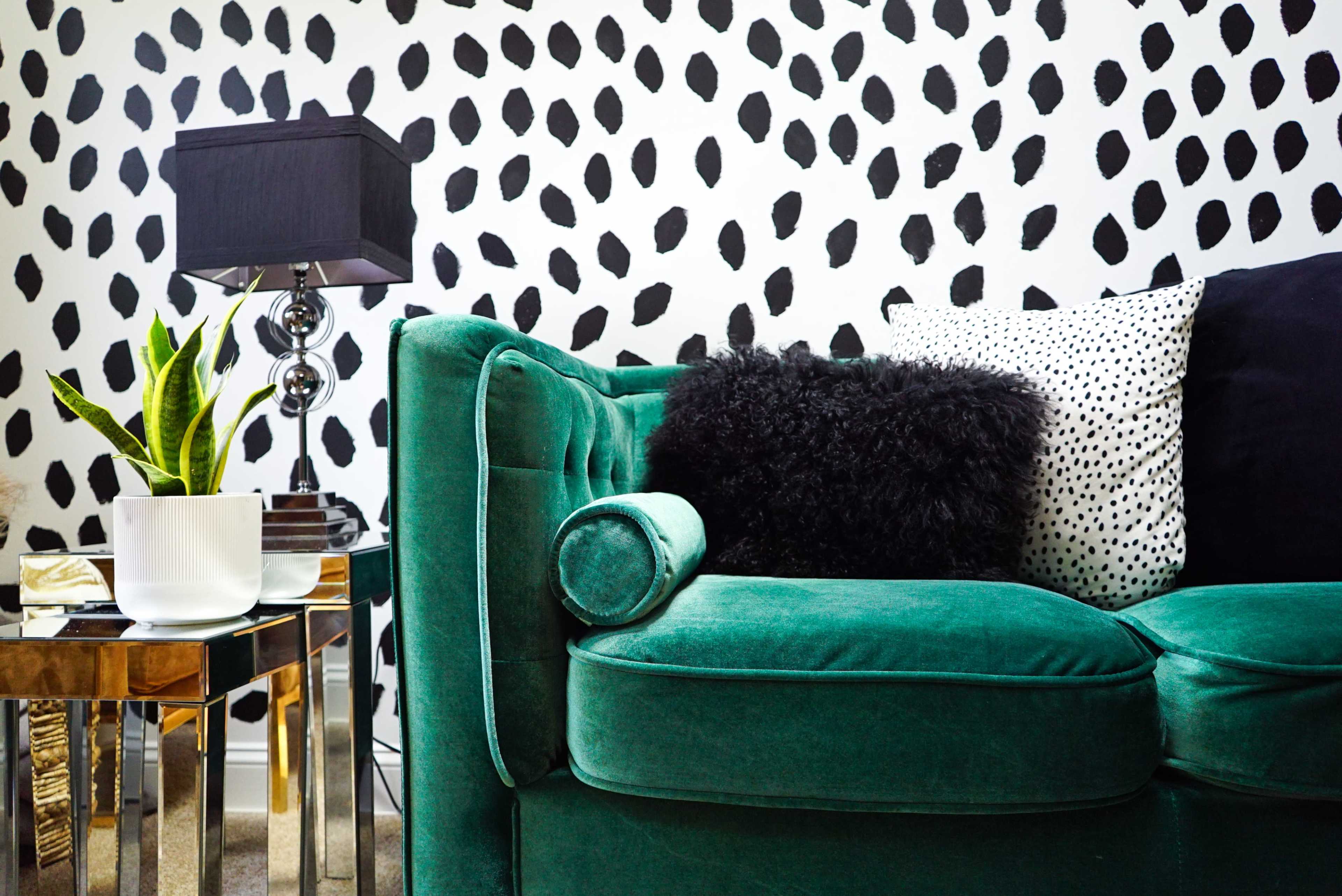 The image features a green velvet couch adorned with various pillows, alongside a side table holding a potted plant and a black lamp, all against a backdrop of a black and white dotted wall.