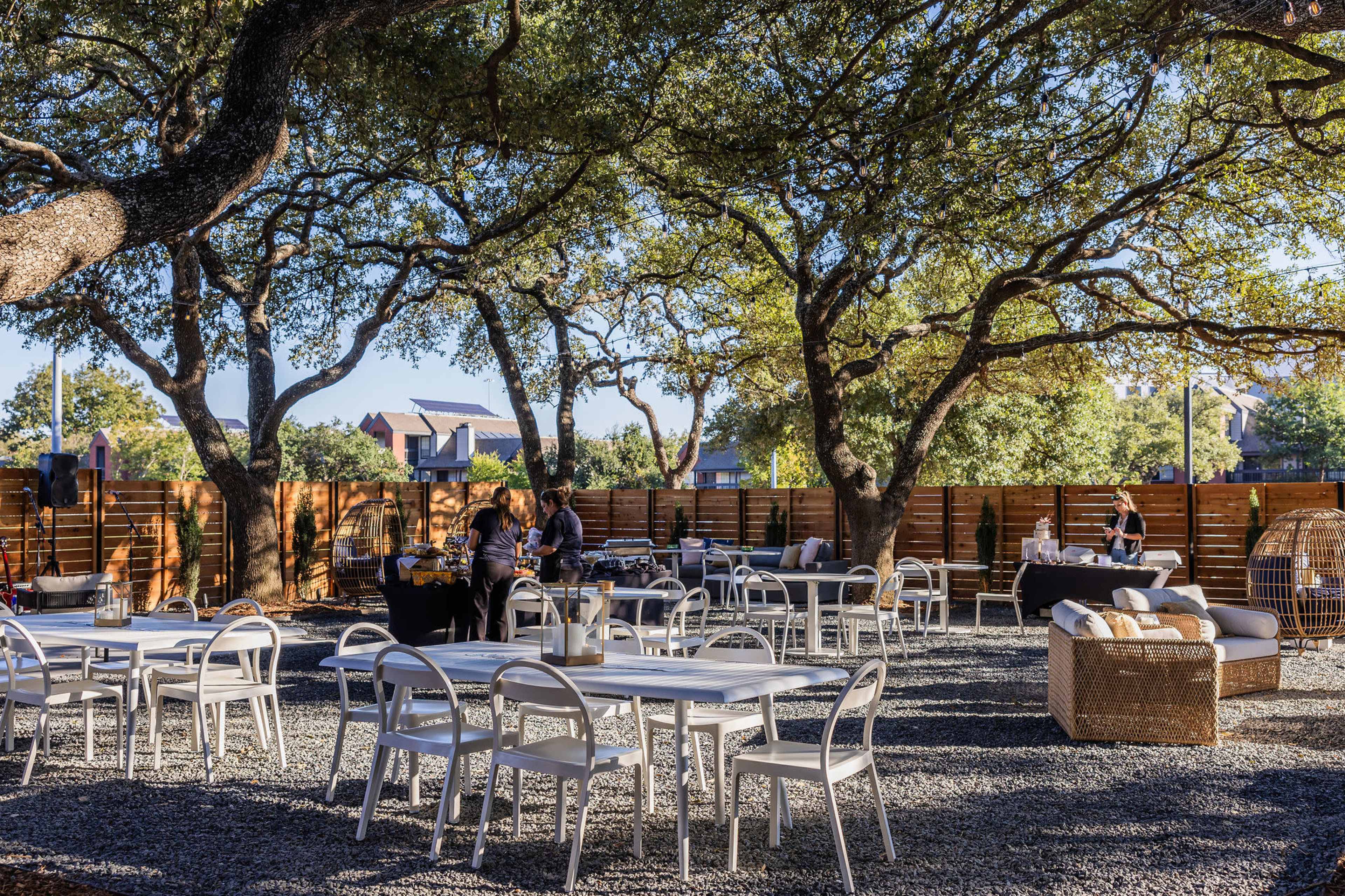 An outdoor dining area features white tables and chairs set on gravel, surrounded by large trees and a wooden fence.