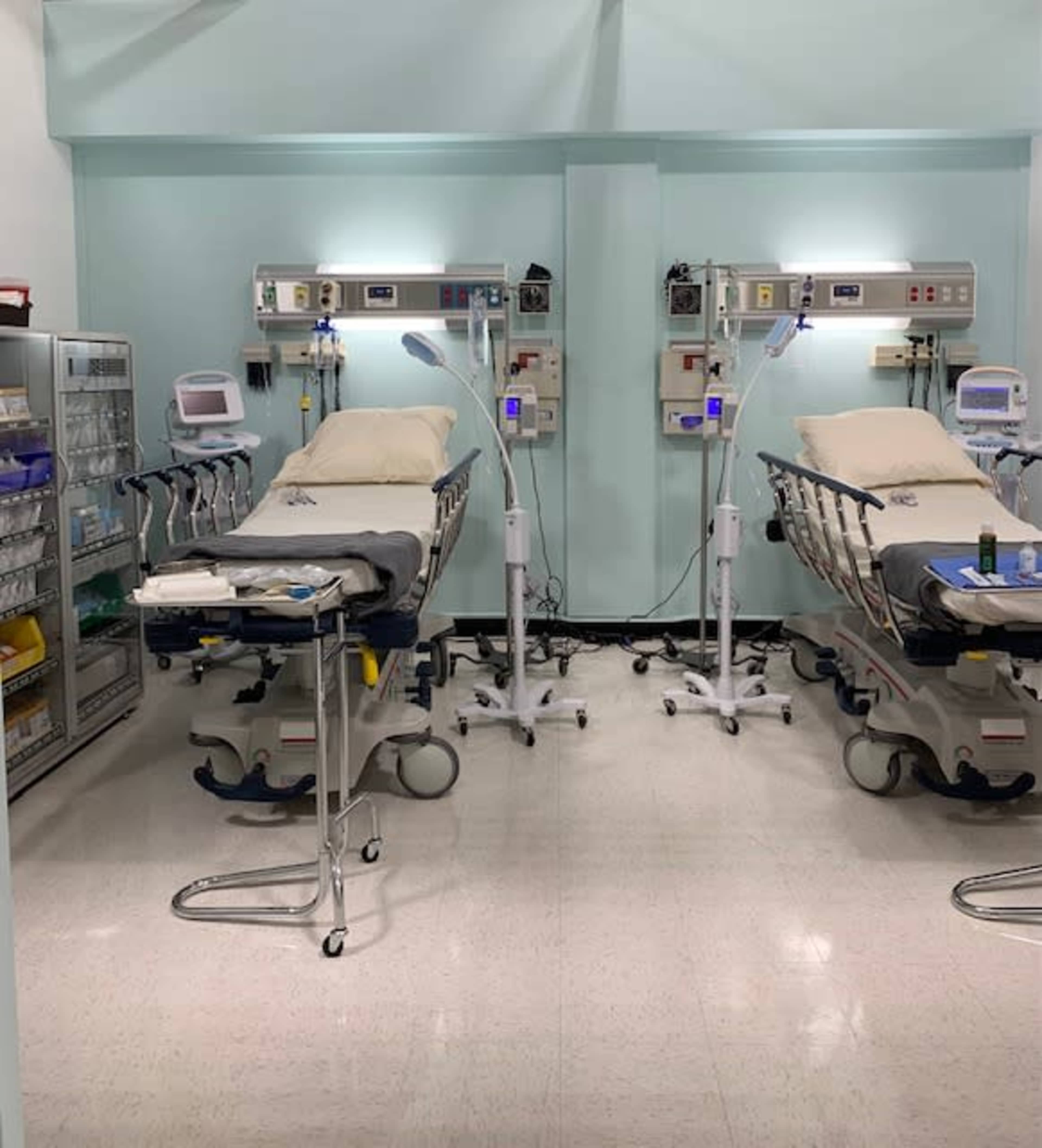 A medical facility with two patient beds, each equipped with electronic monitors and medical equipment, positioned beside a storage unit filled with supplies.