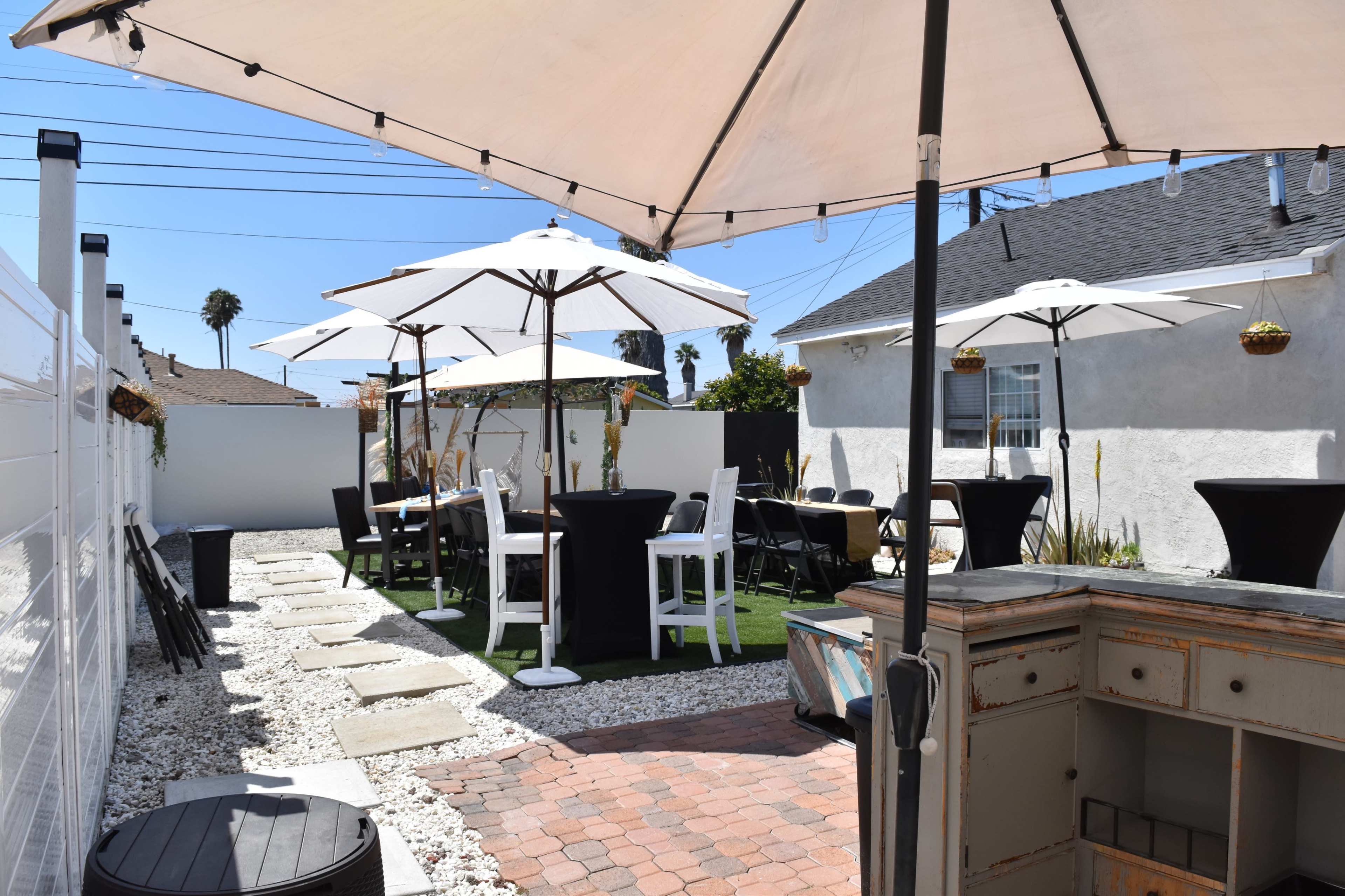 An outdoor patio area with white umbrellas over tables and chairs arranged on a lawn surrounded by decorative stone pathways.