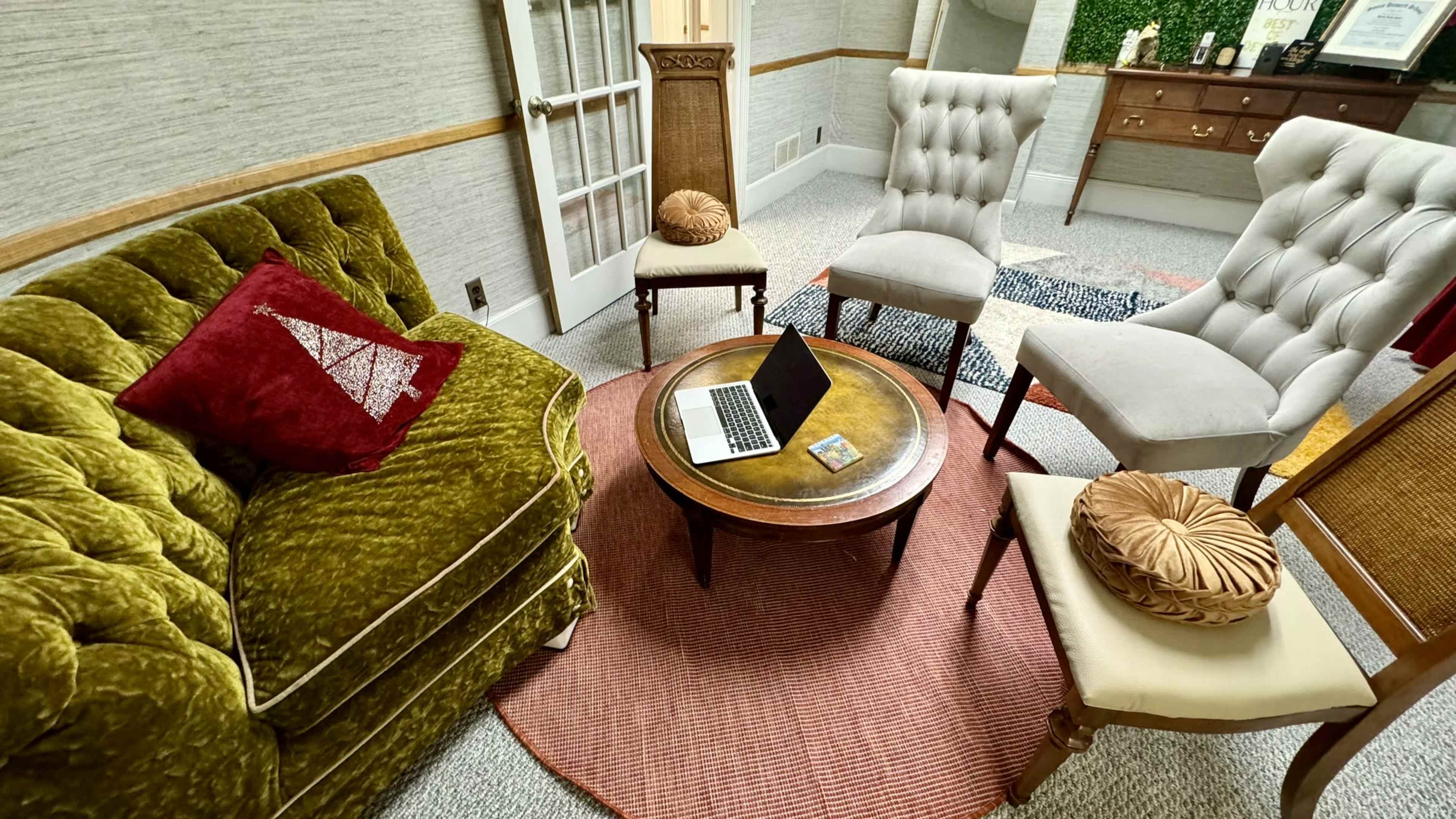 A cozy sitting area features a green velvet sofa, a round coffee table with a laptop, and several upholstered chairs arranged on a patterned rug.