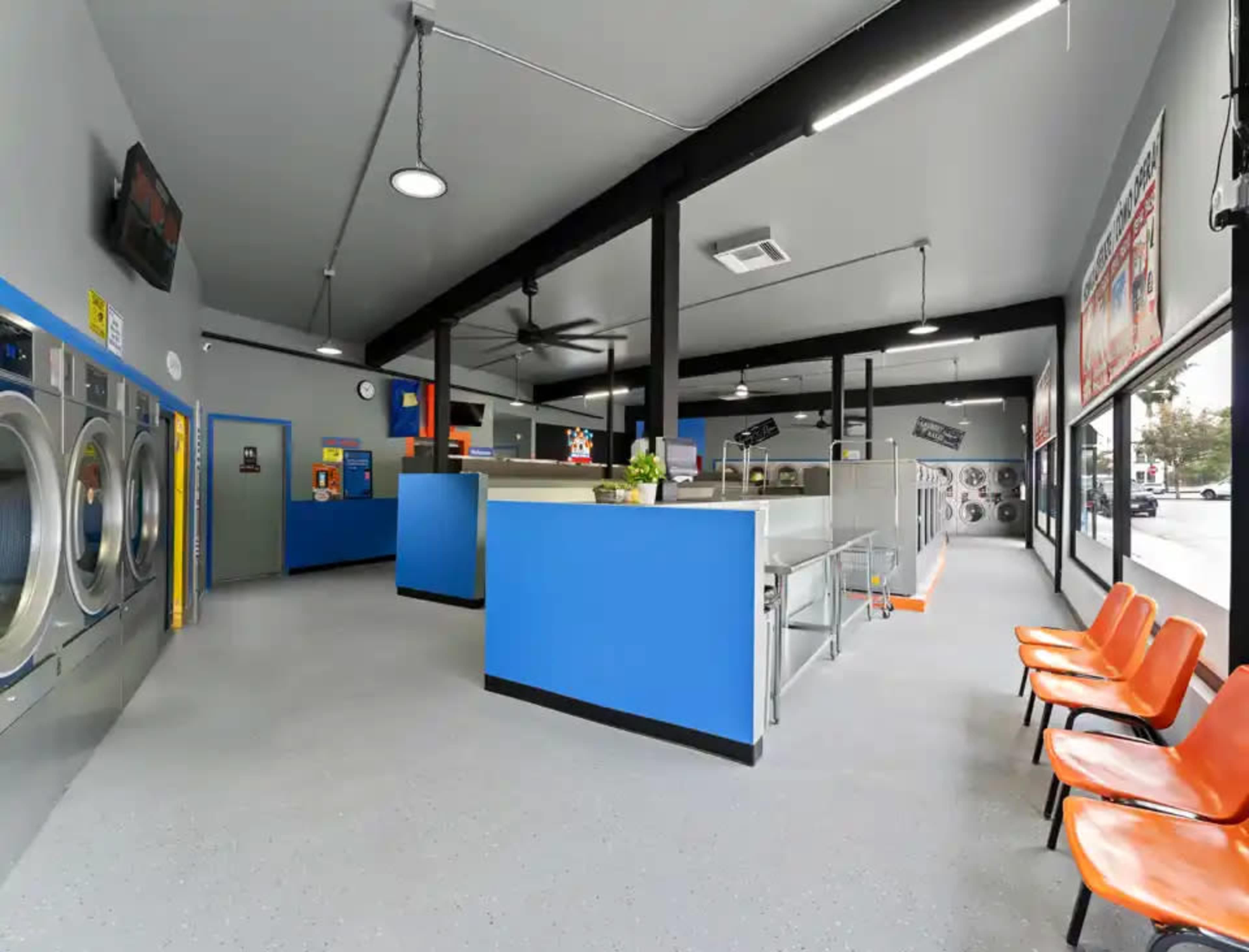 The image shows the interior of a laundromat with washing machines on one side, orange chairs arranged in front, and a bright, open layout featuring blue and gray decor.