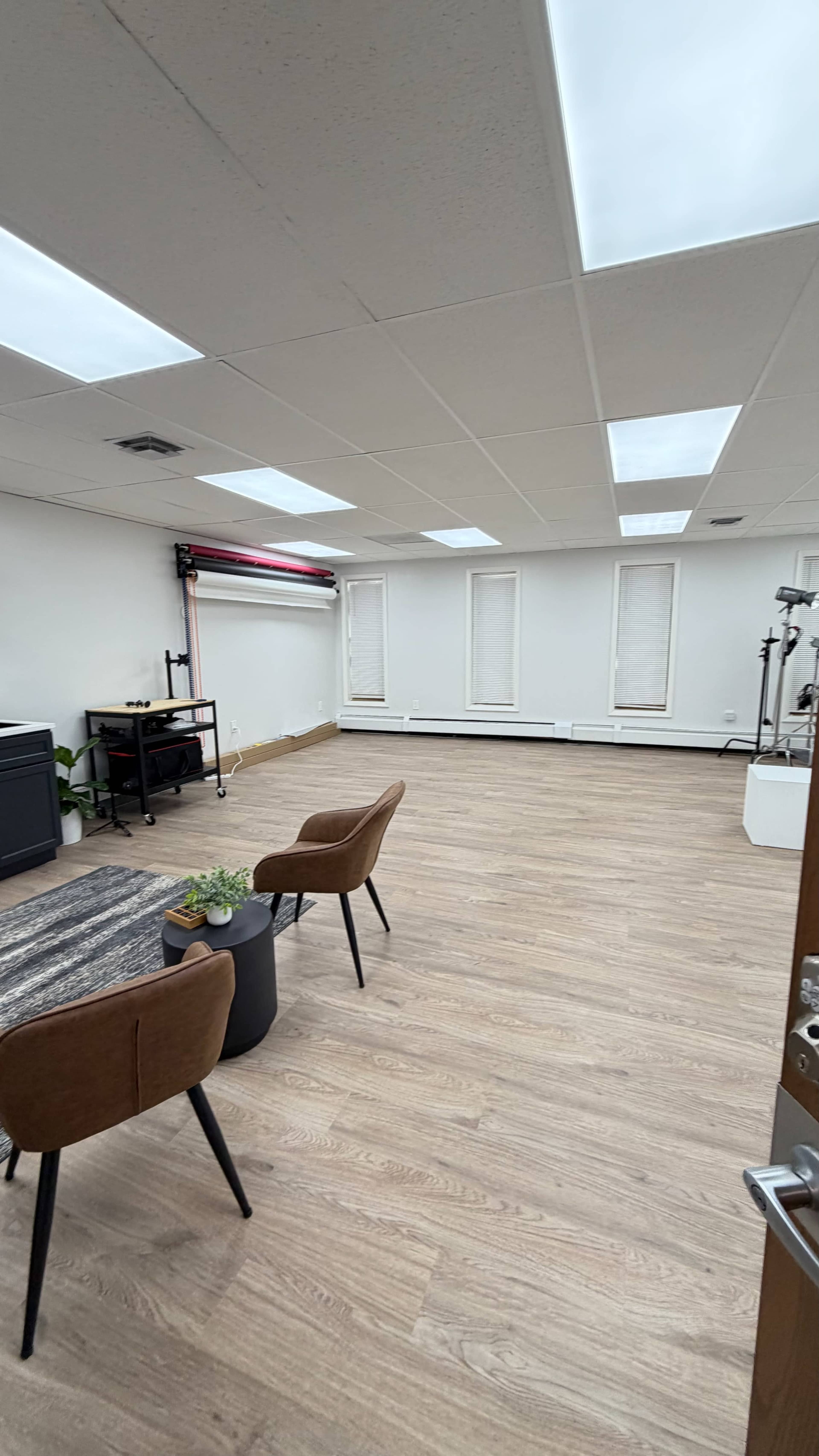 The image shows a spacious, empty room with hardwood floors, featuring two chairs, a small table, and various studio equipment along the walls.
