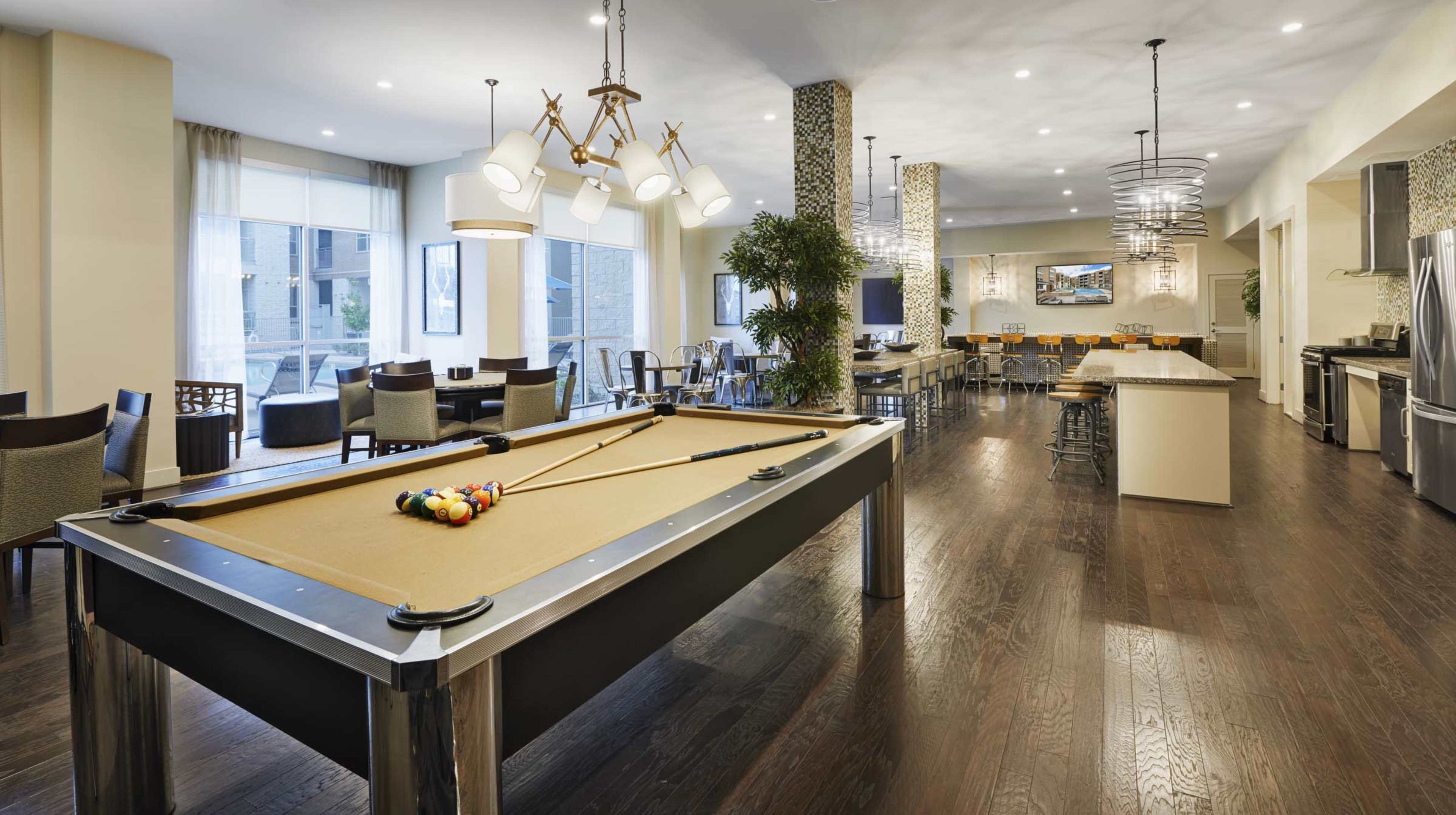 The image shows a modern recreational room featuring a pool table, seating areas, and a bar counter with kitchen appliances in an open layout.