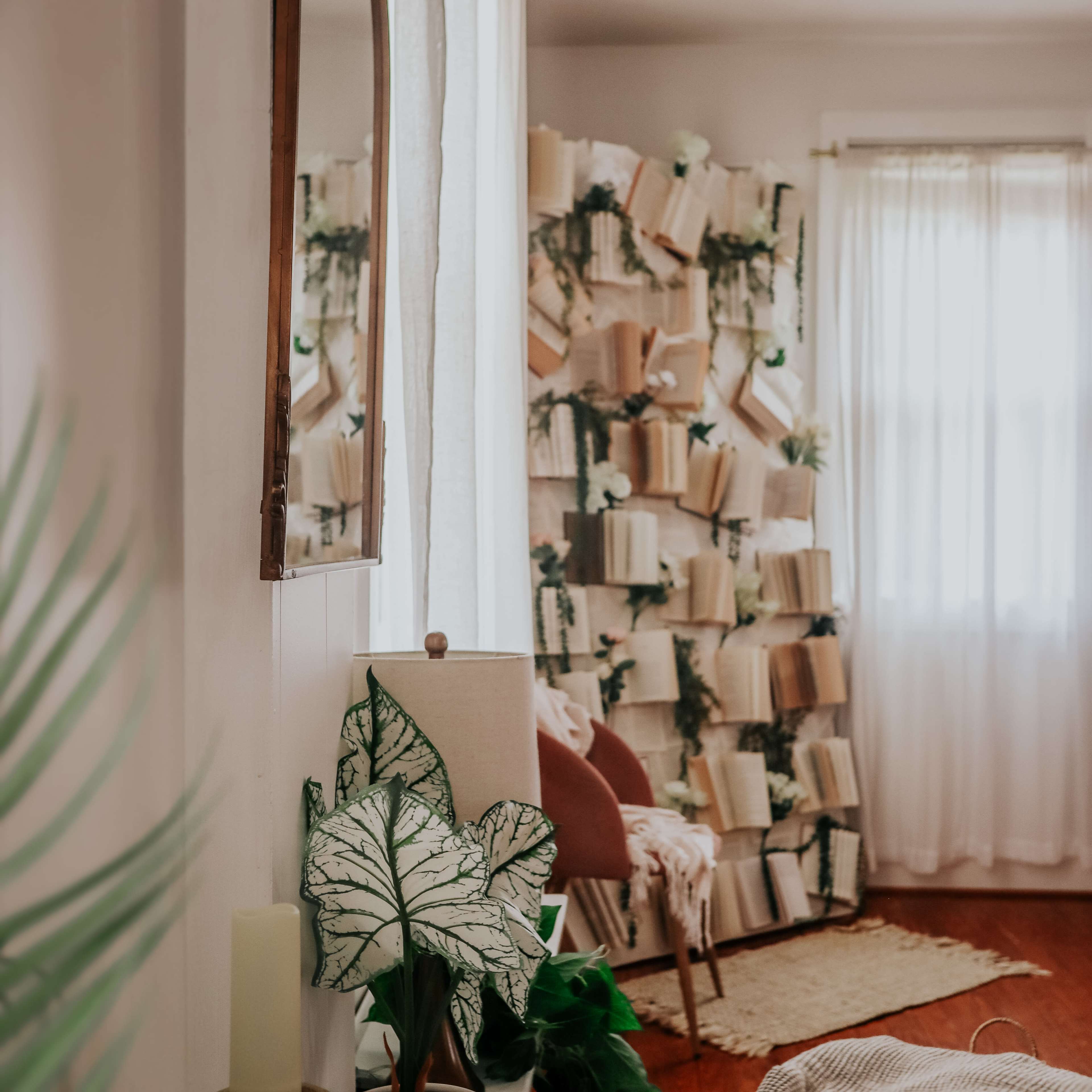 A cozy room features a mirror, a lamp with a leafy plant, and a decorative wall covered in arranged books and greenery.