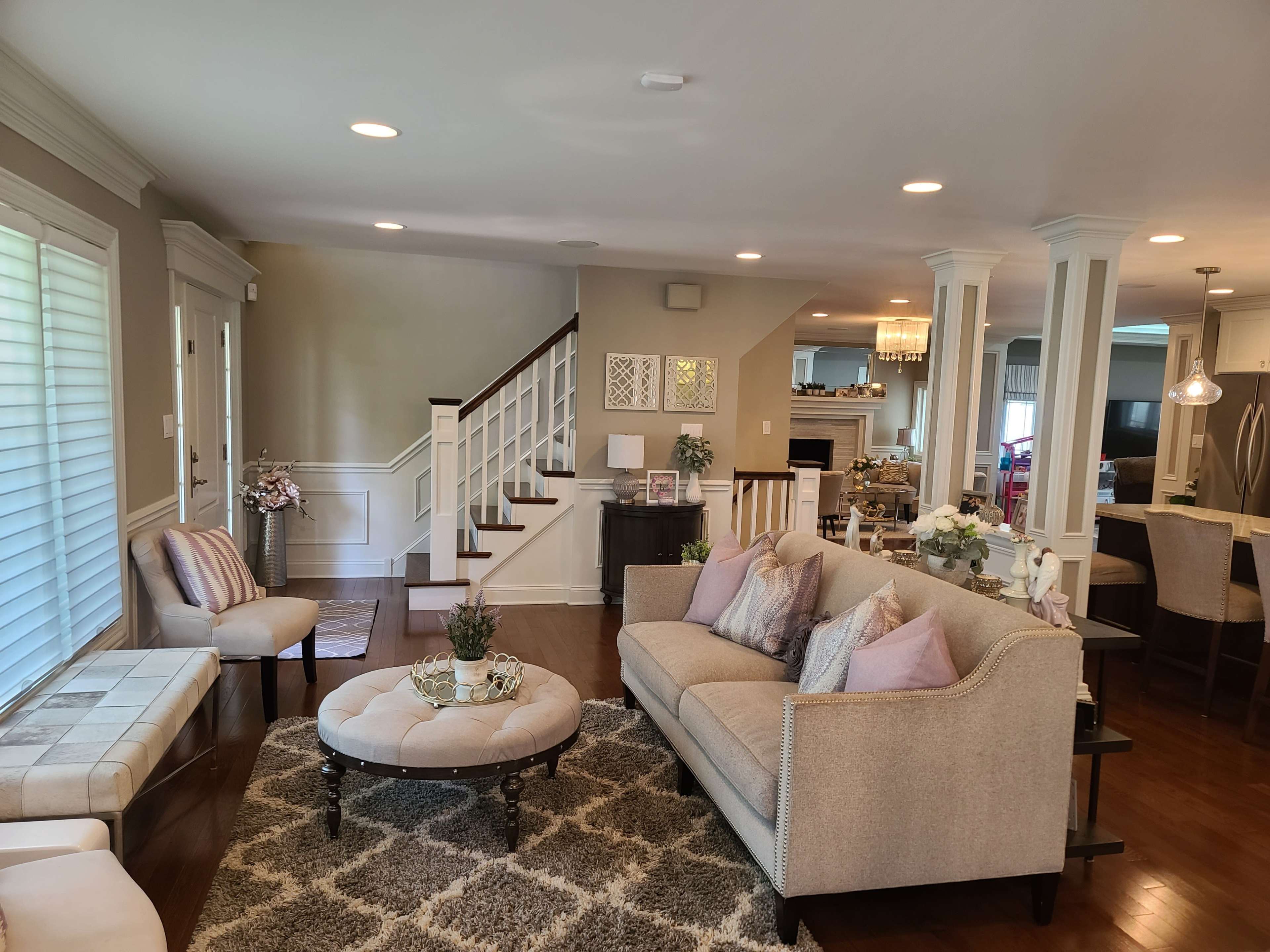 The image shows a spacious living room with a light-colored sofa, an ottoman, and a staircase leading to the upper floor.