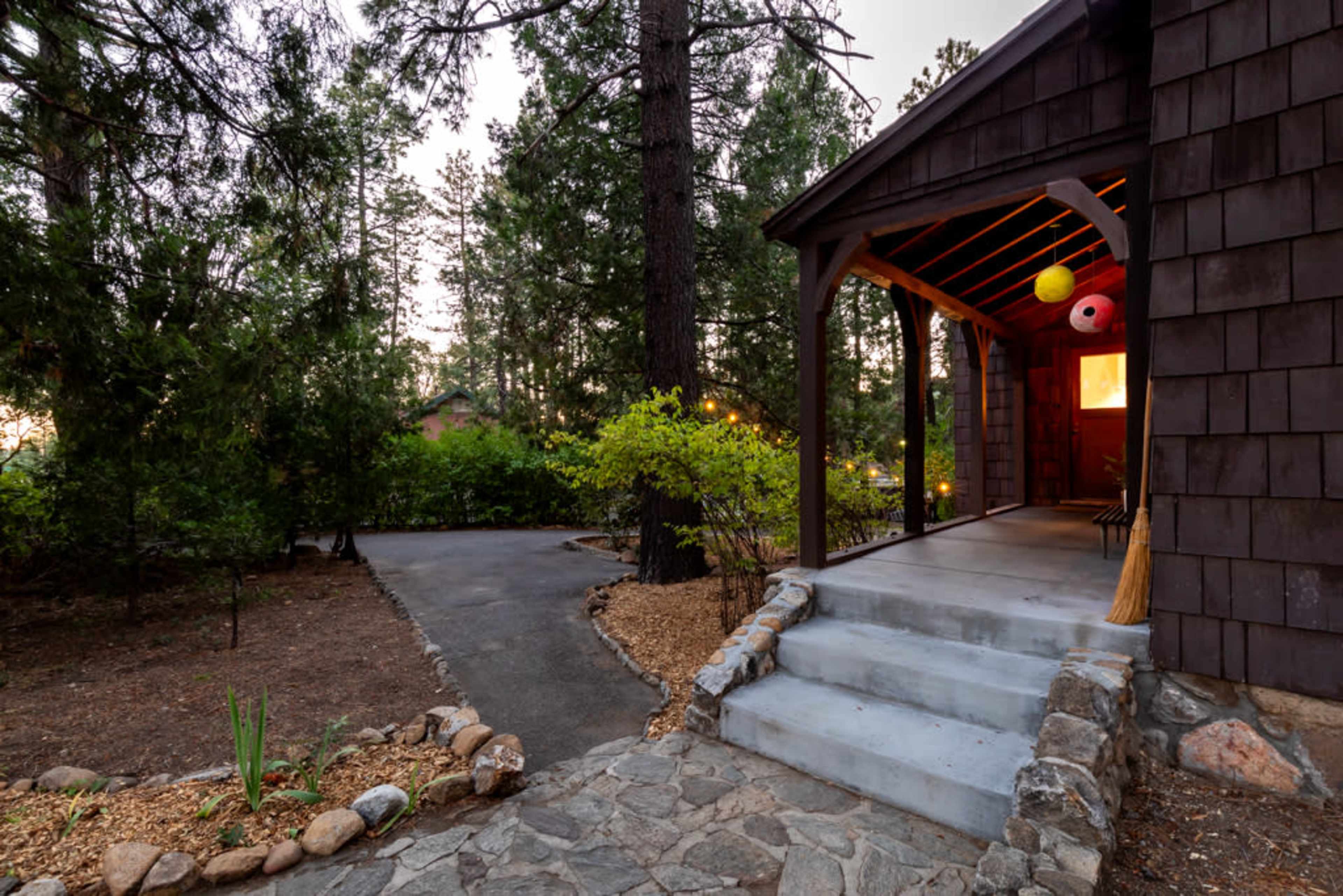 The image shows a path leading to the entrance of a wooden cabin surrounded by tall trees and landscaped shrubs.