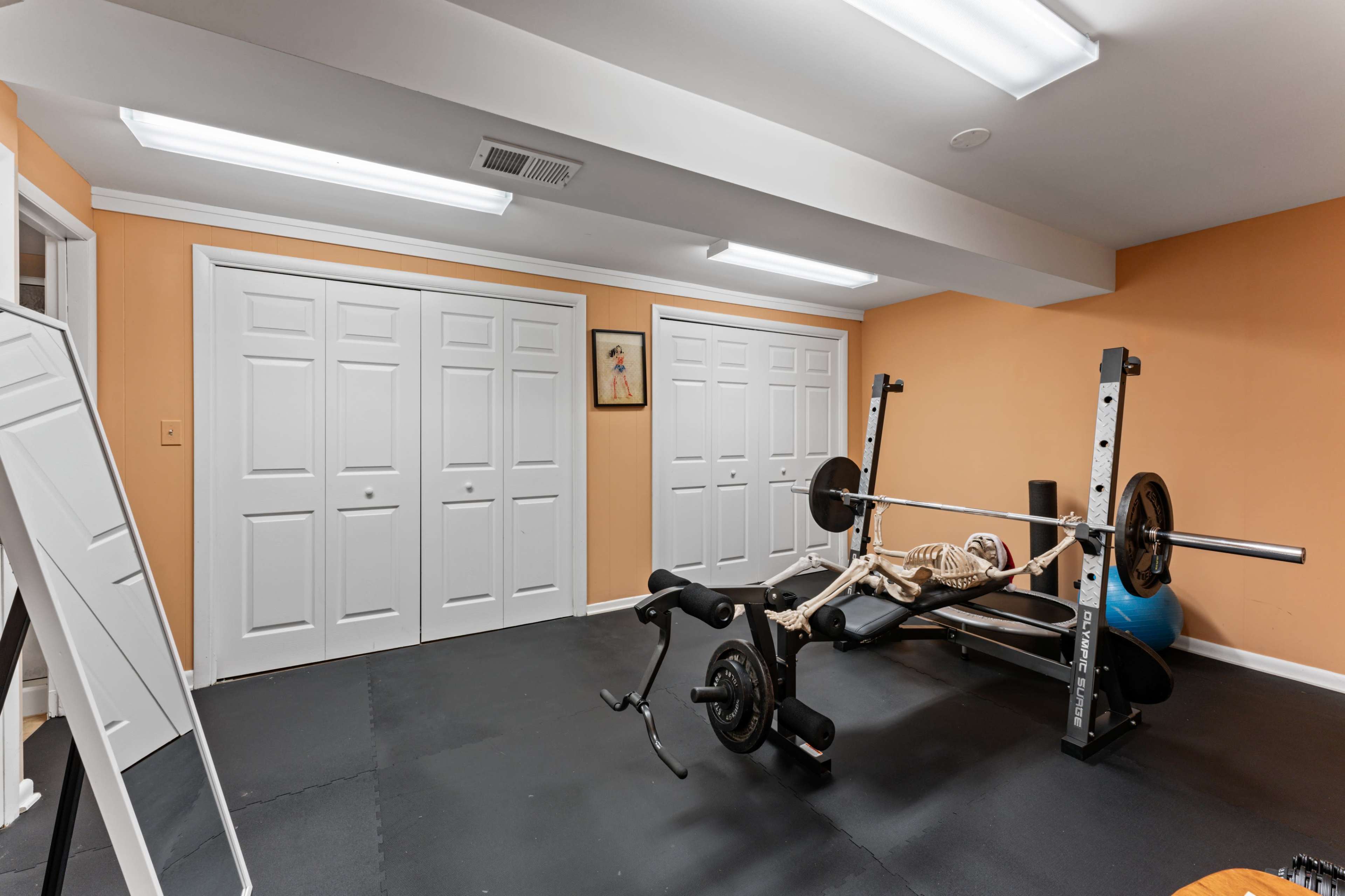 The image shows a home gym with a weight bench, barbell, and a skeleton figure positioned on the bench, situated against orange walls and featuring mirrored and closed closet doors.