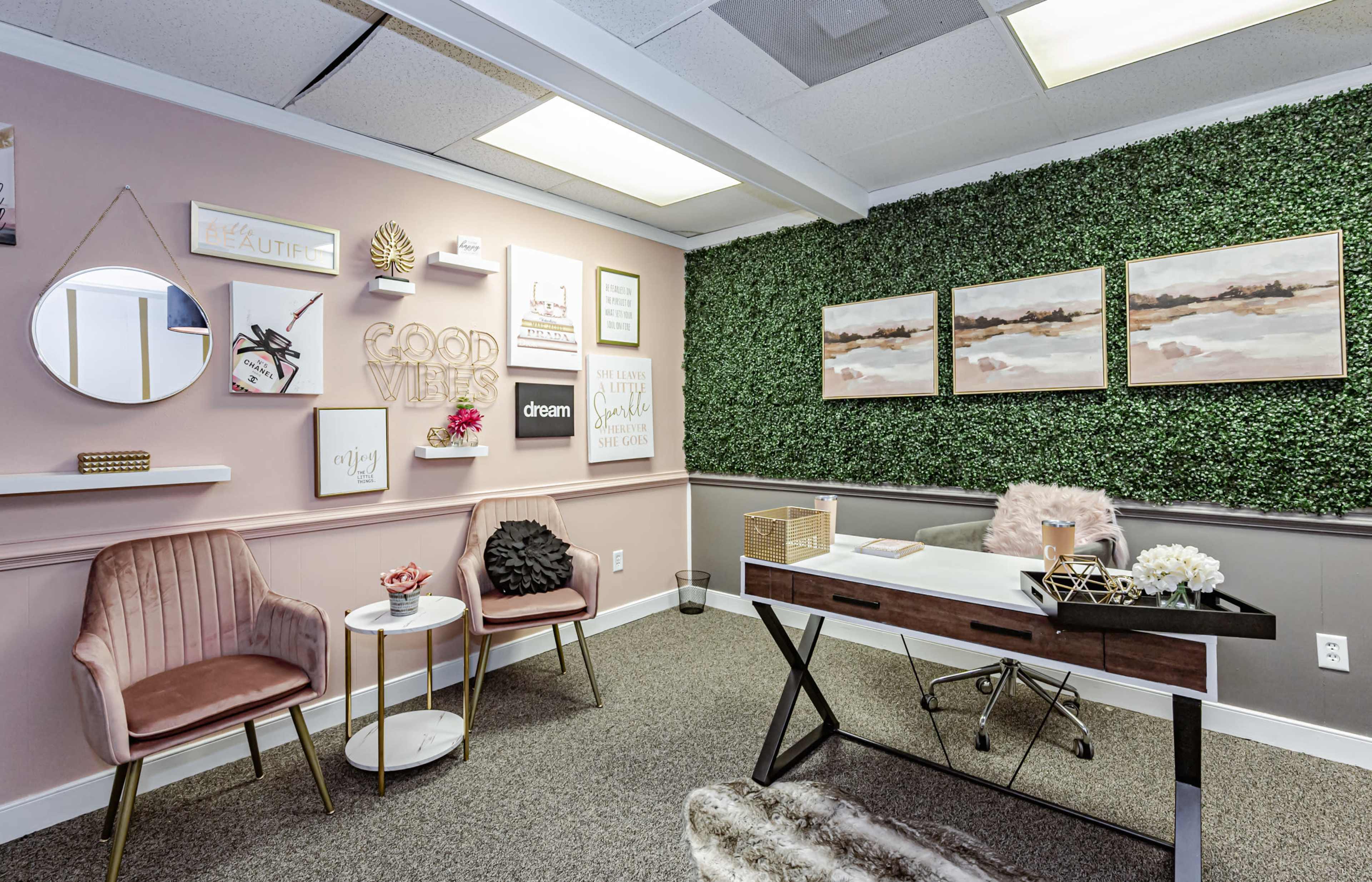 A stylish office space featuring a desk, decorative wall art, and pink accent chairs against a backdrop of green plants.