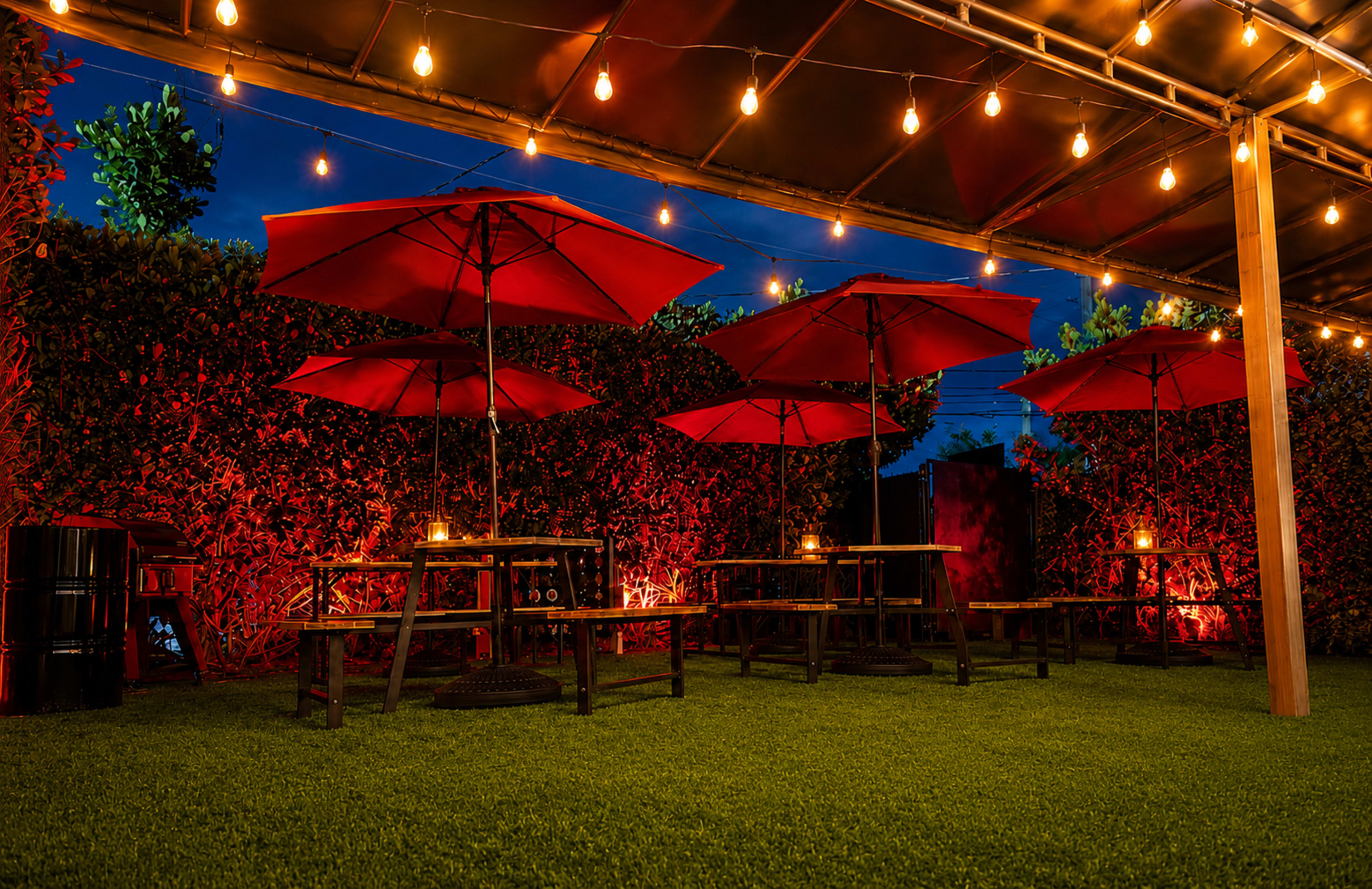 The image shows a patio area with several tables and red umbrellas under warm string lights, surrounded by greenery.