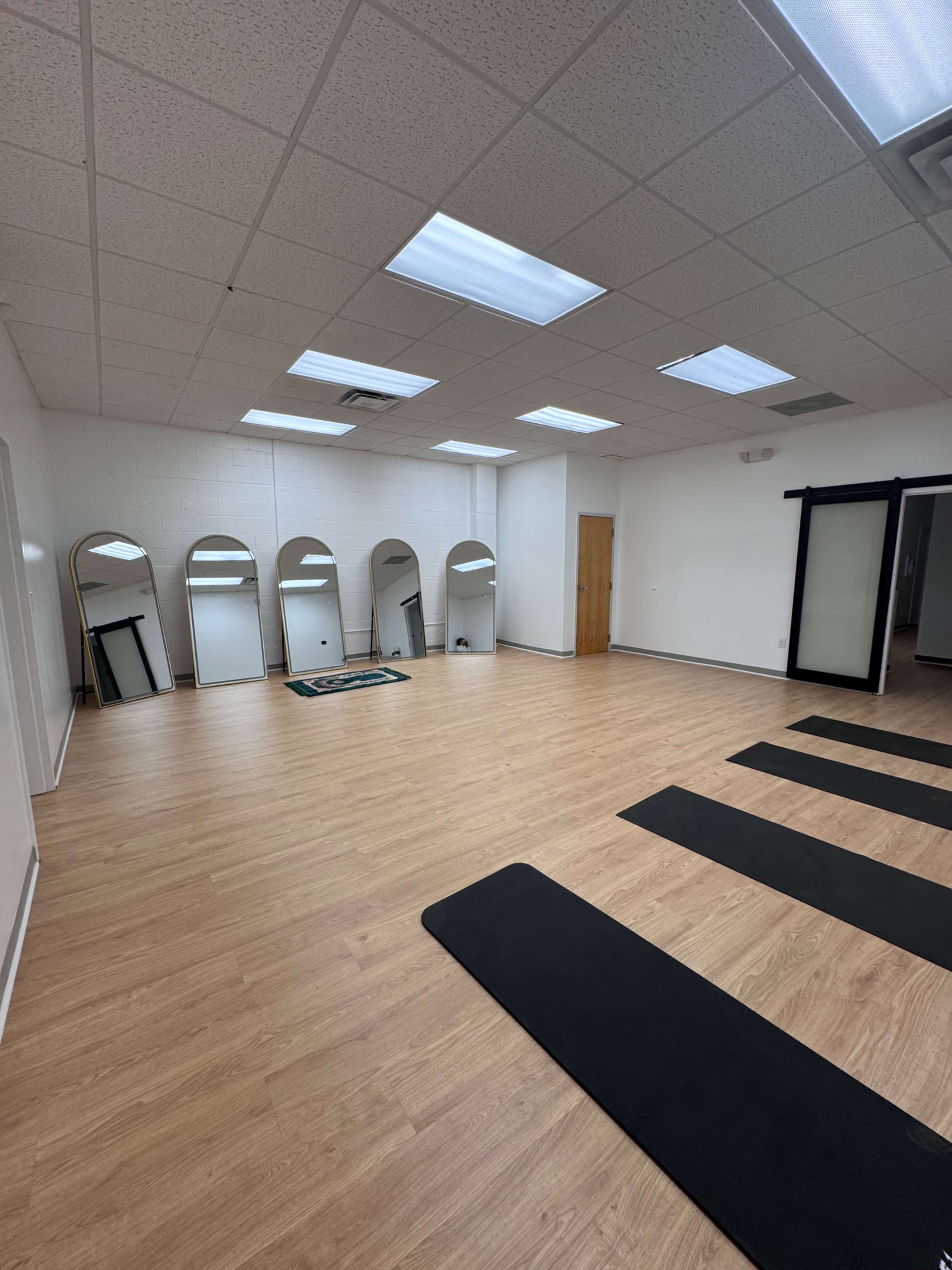 The image shows an empty studio space with wooden flooring, yoga mats aligned on the floor, and several arched mirrors on the wall.