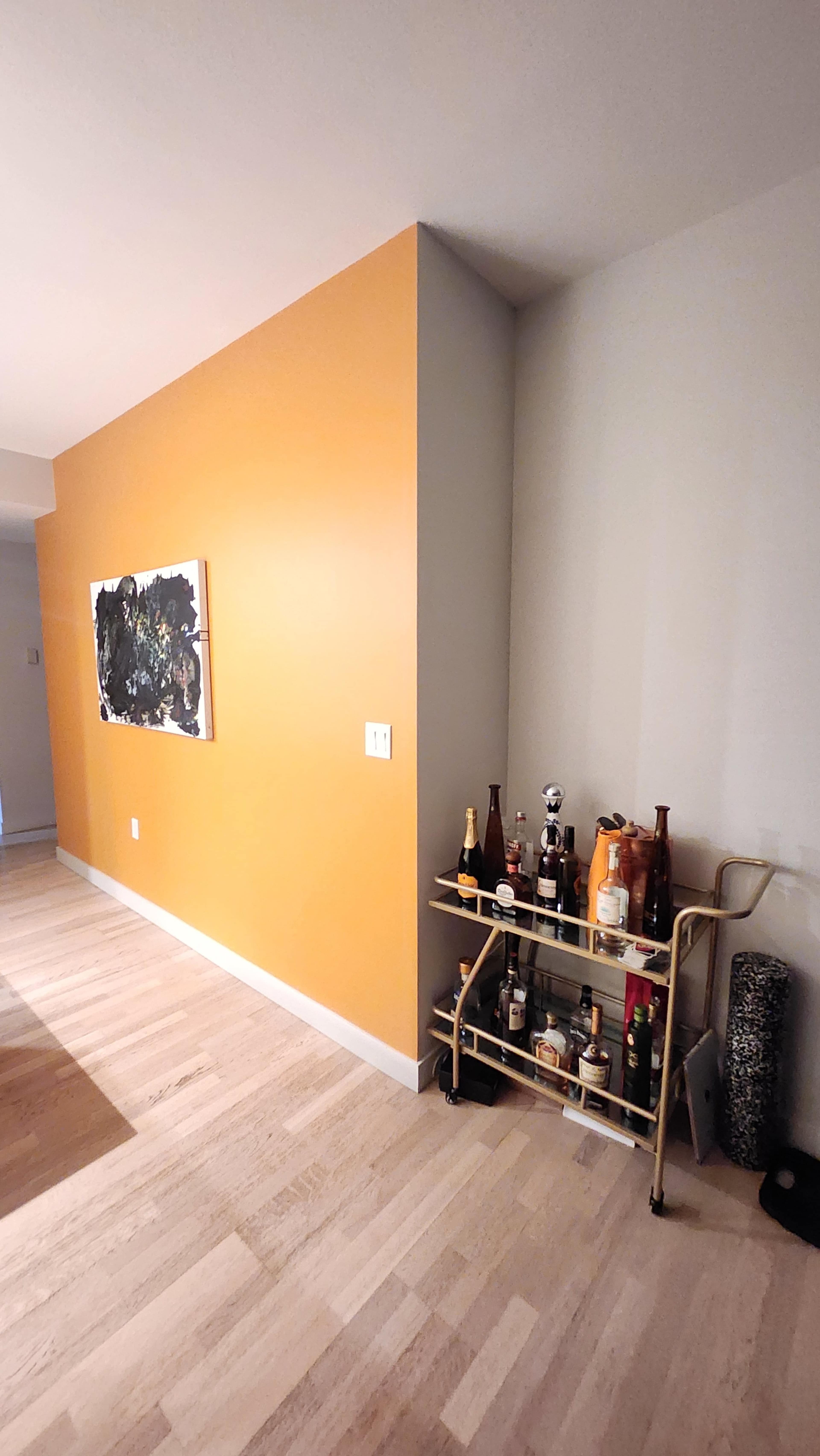 A bright orange wall meets a light gray wall with a bar cart displaying various bottles in the corner of a room.