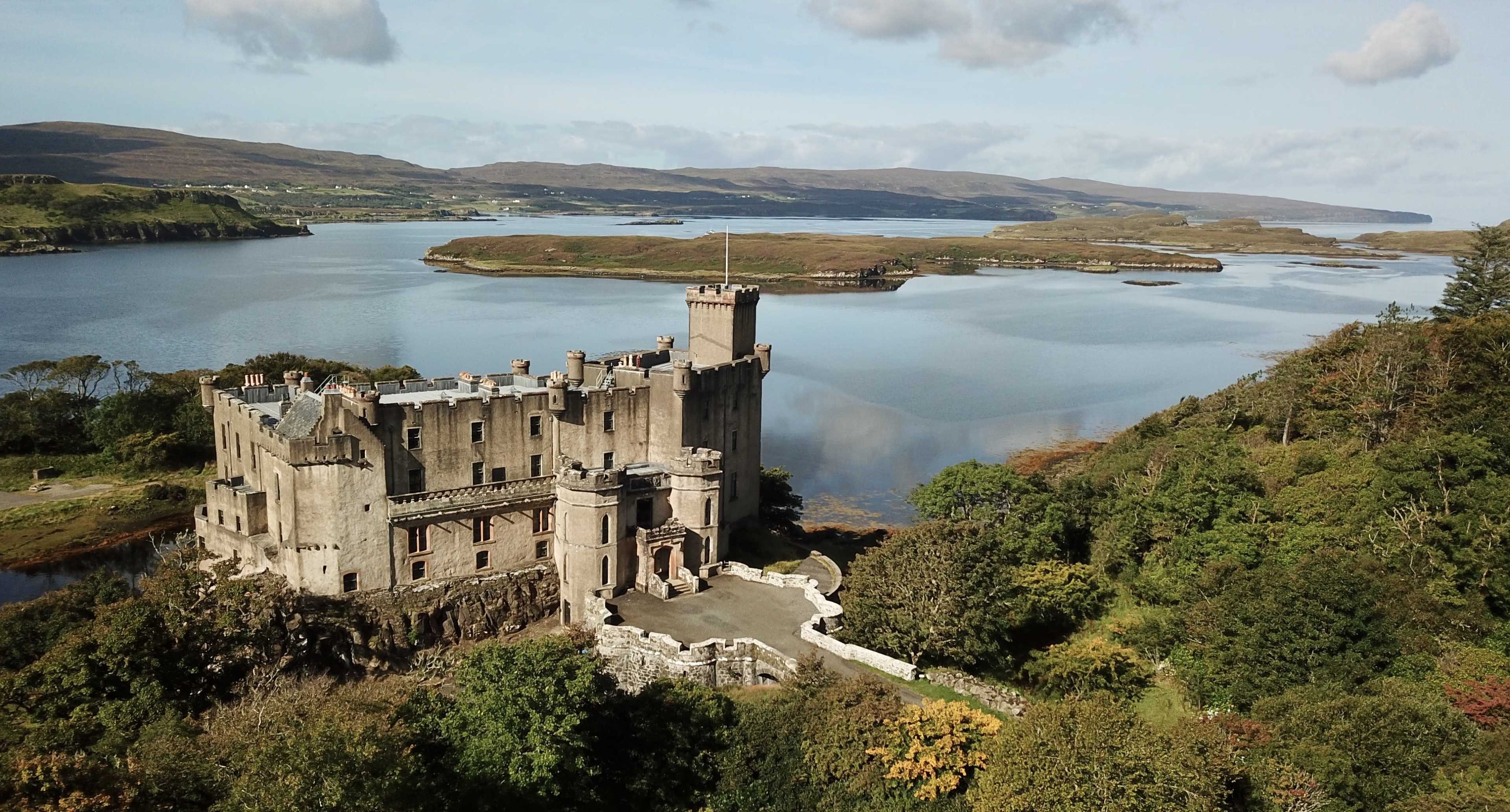 The image shows a stone castle situated on the edge of a body of water, surrounded by green trees and hills in the background.