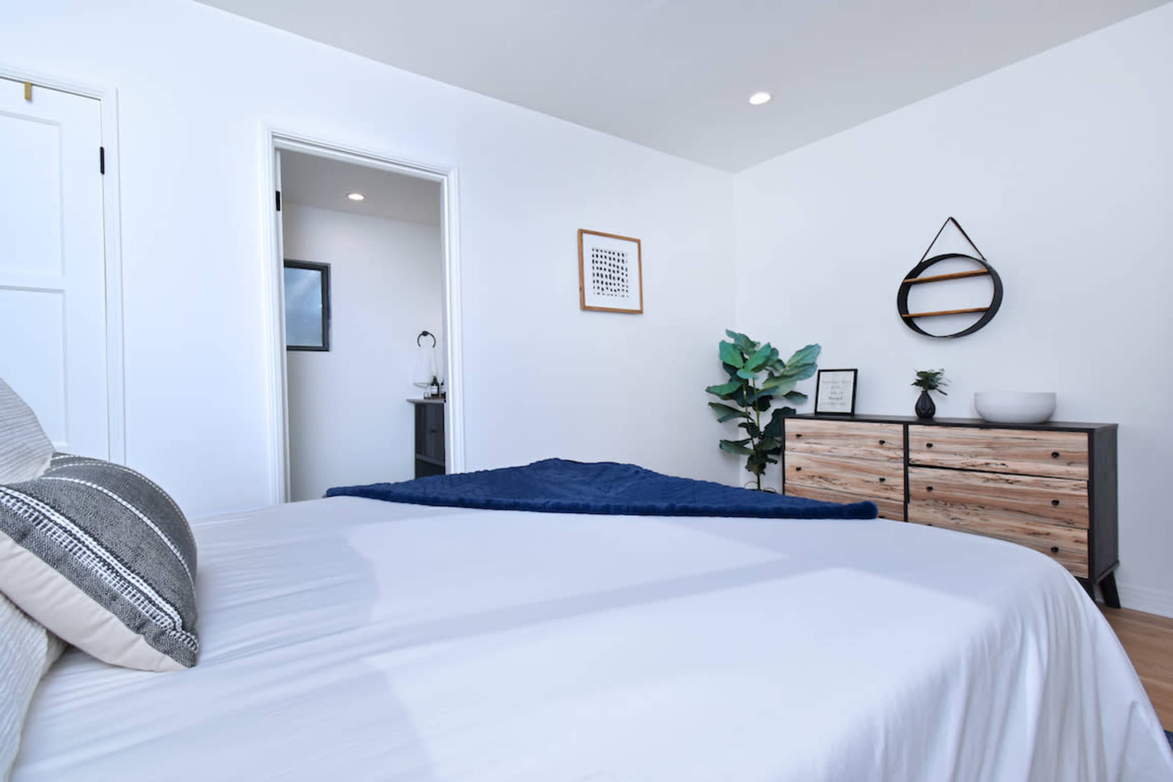 The image shows a well-lit bedroom featuring a large bed, a dresser, and a plant, with a doorway leading to another room in the background.