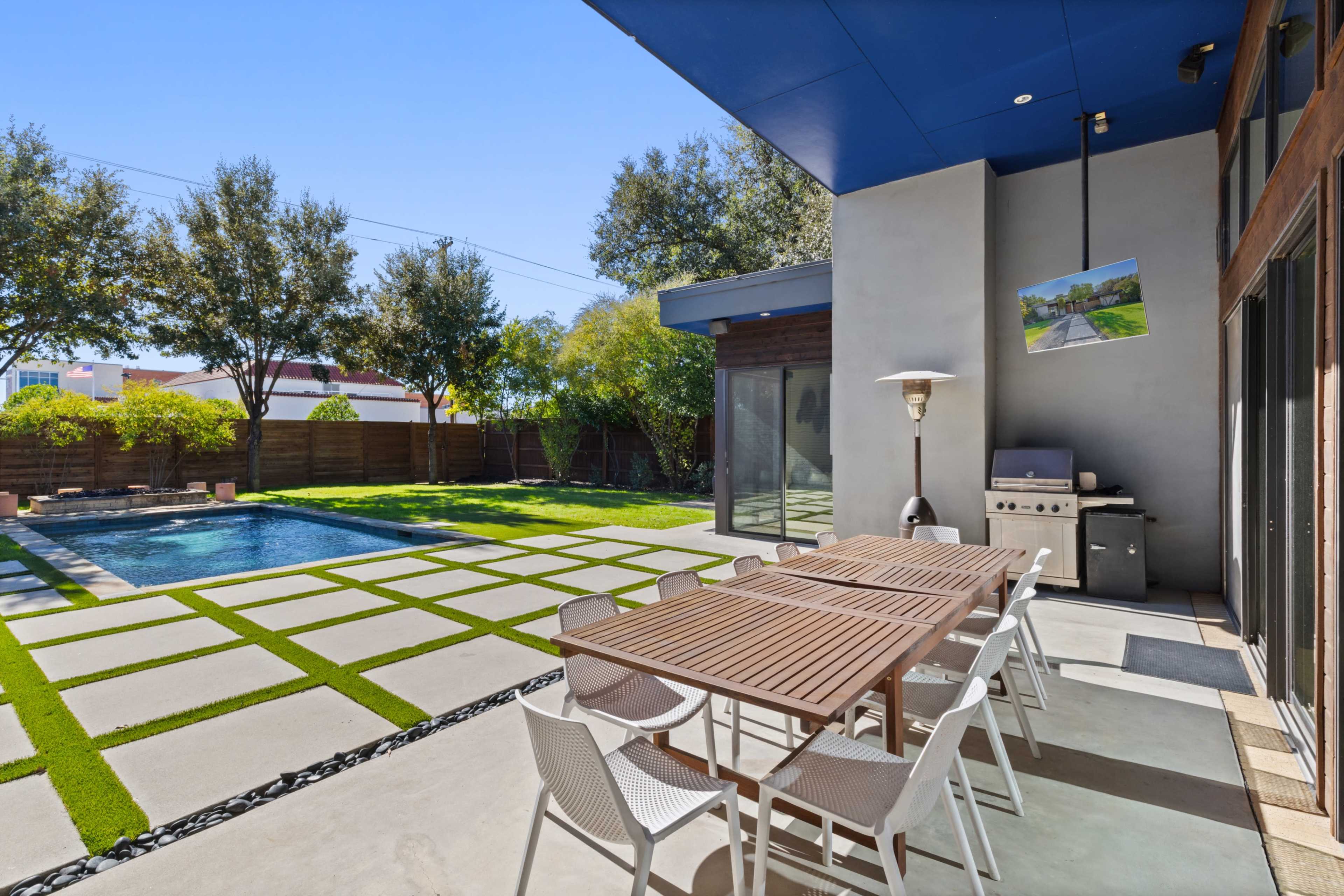 The image shows a modern outdoor patio area with a dining table and chairs, a grill, and a swimming pool surrounded by green landscaping.