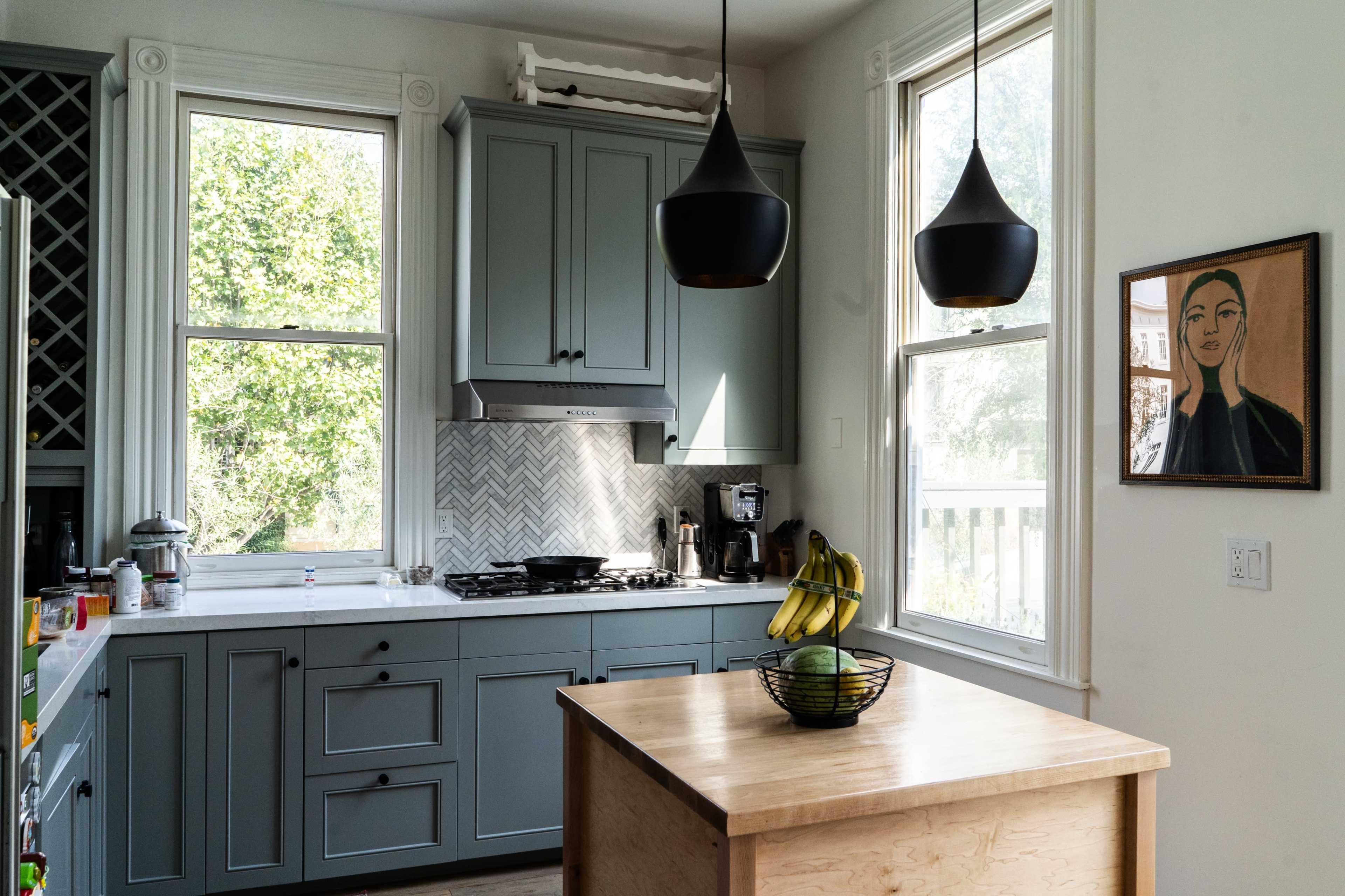 A modern kitchen features light blue cabinets, a central wooden island with a bowl of bananas, and large windows allowing natural light, framed by two black pendant lights.