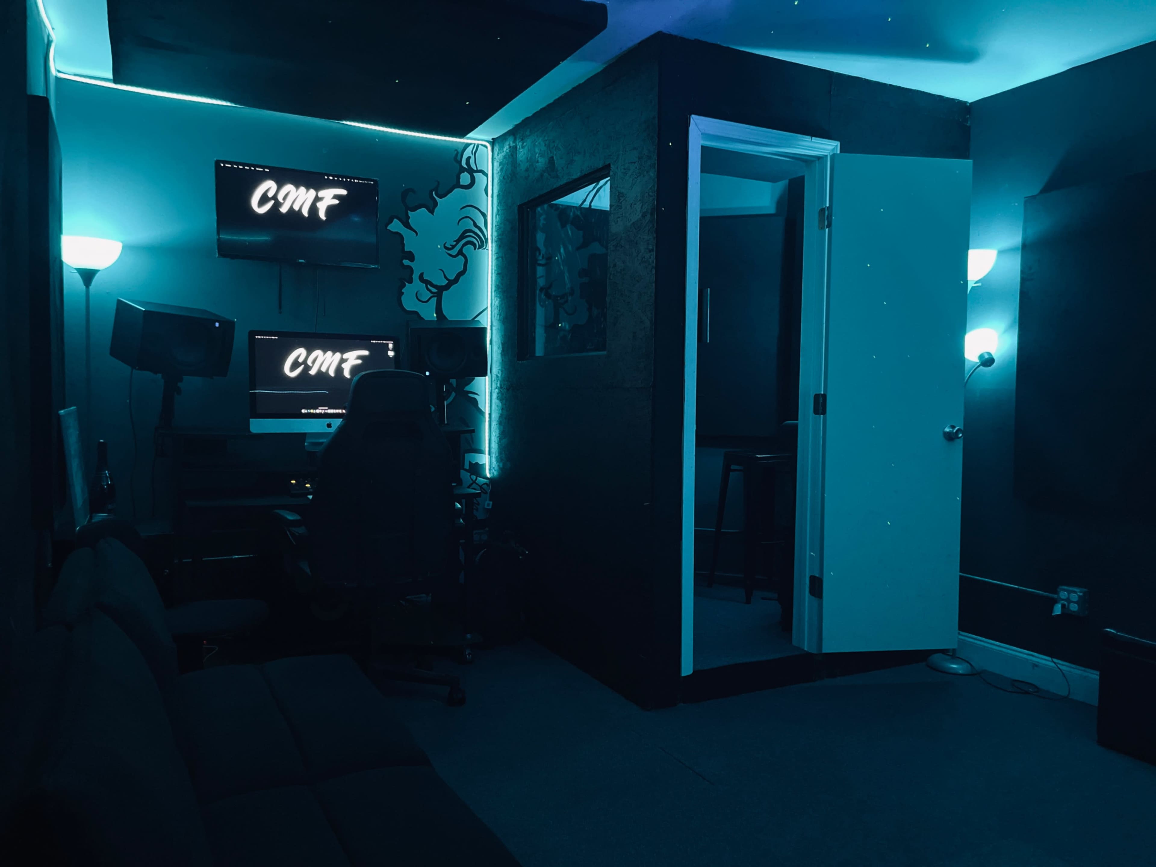 The image shows a dimly lit sound studio with blue lighting, featuring a computer setup, speakers, a recording booth, and a seating area.