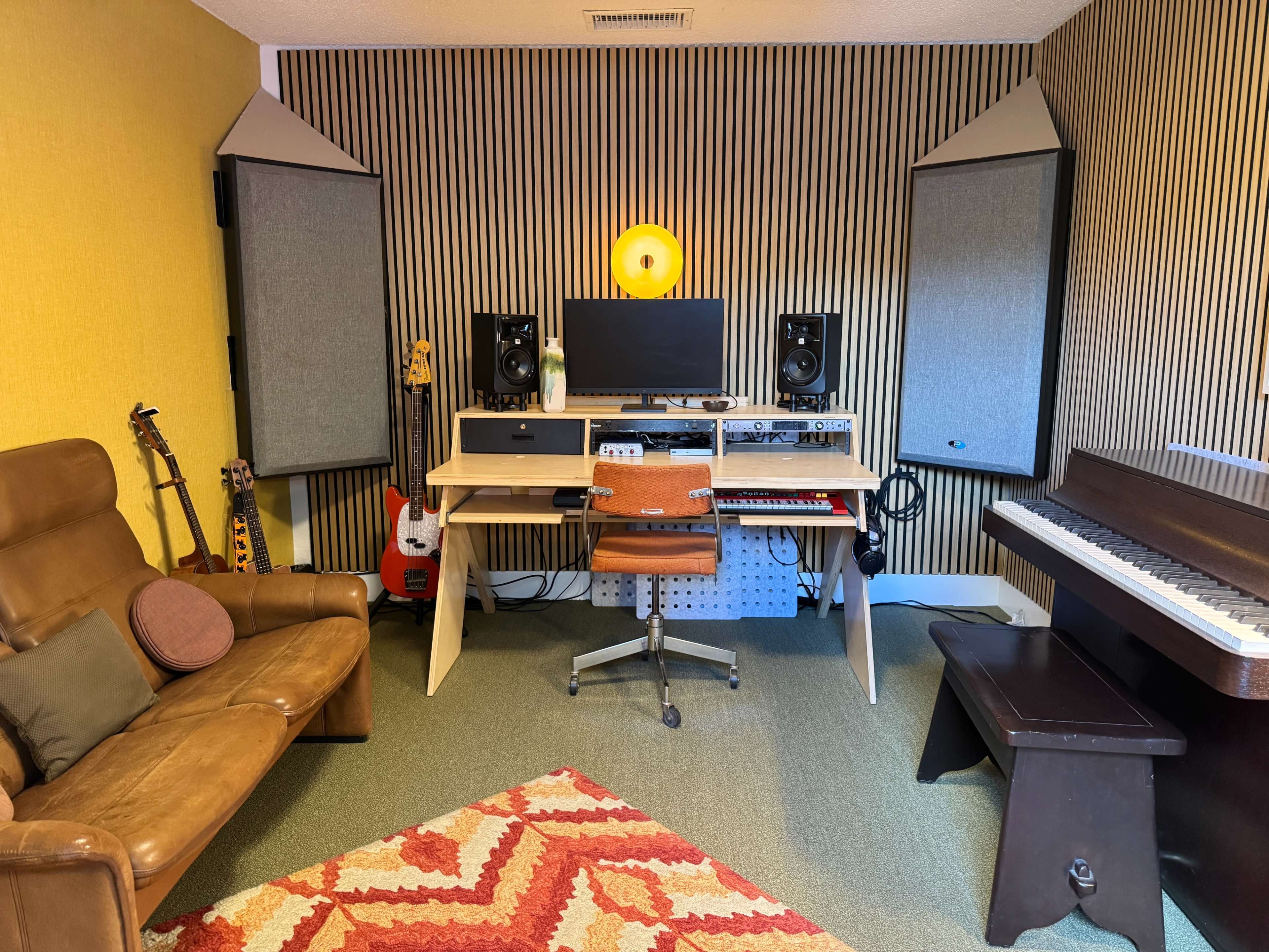 The image features a music studio with a brown leather couch, a desk with a computer and audio equipment, two large speakers, a piano, and several musical instruments including guitars.