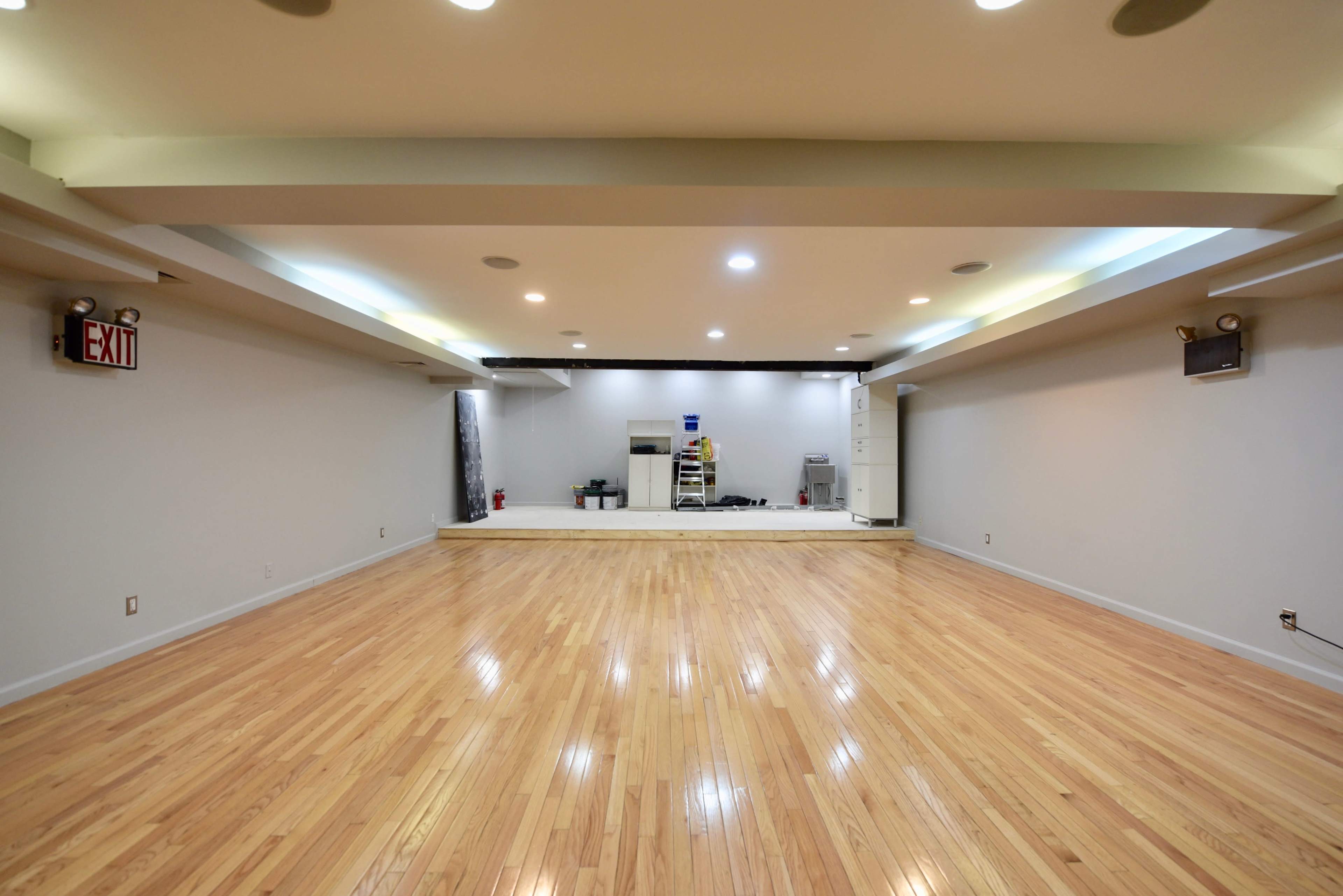 A spacious room with polished wooden floors and a raised platform in the center, featuring a neutral-colored wall and ceiling lights.
