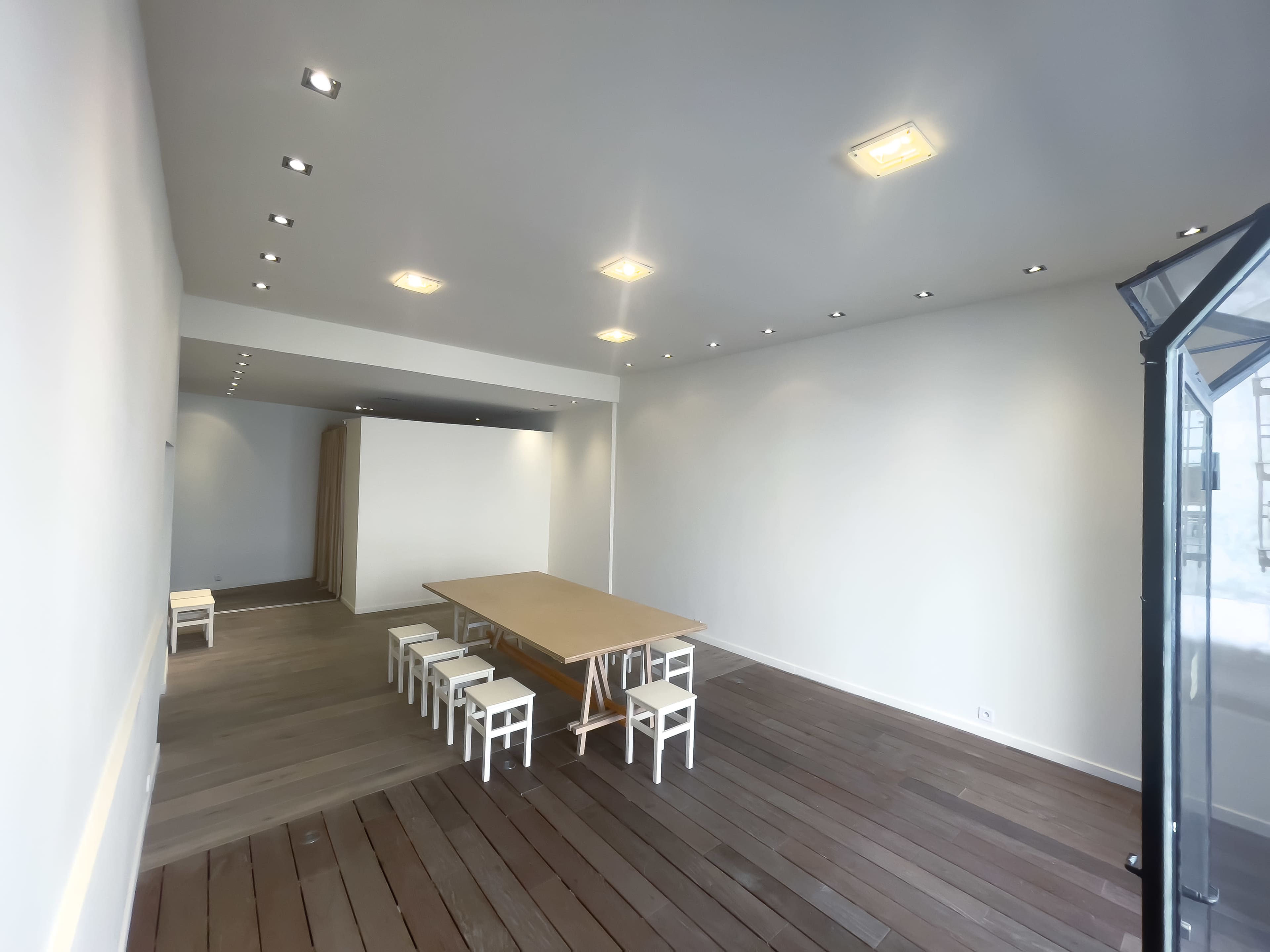 The image shows a spacious, minimalist room with wooden flooring, a large table at the center, and several white chairs arranged neatly around it.