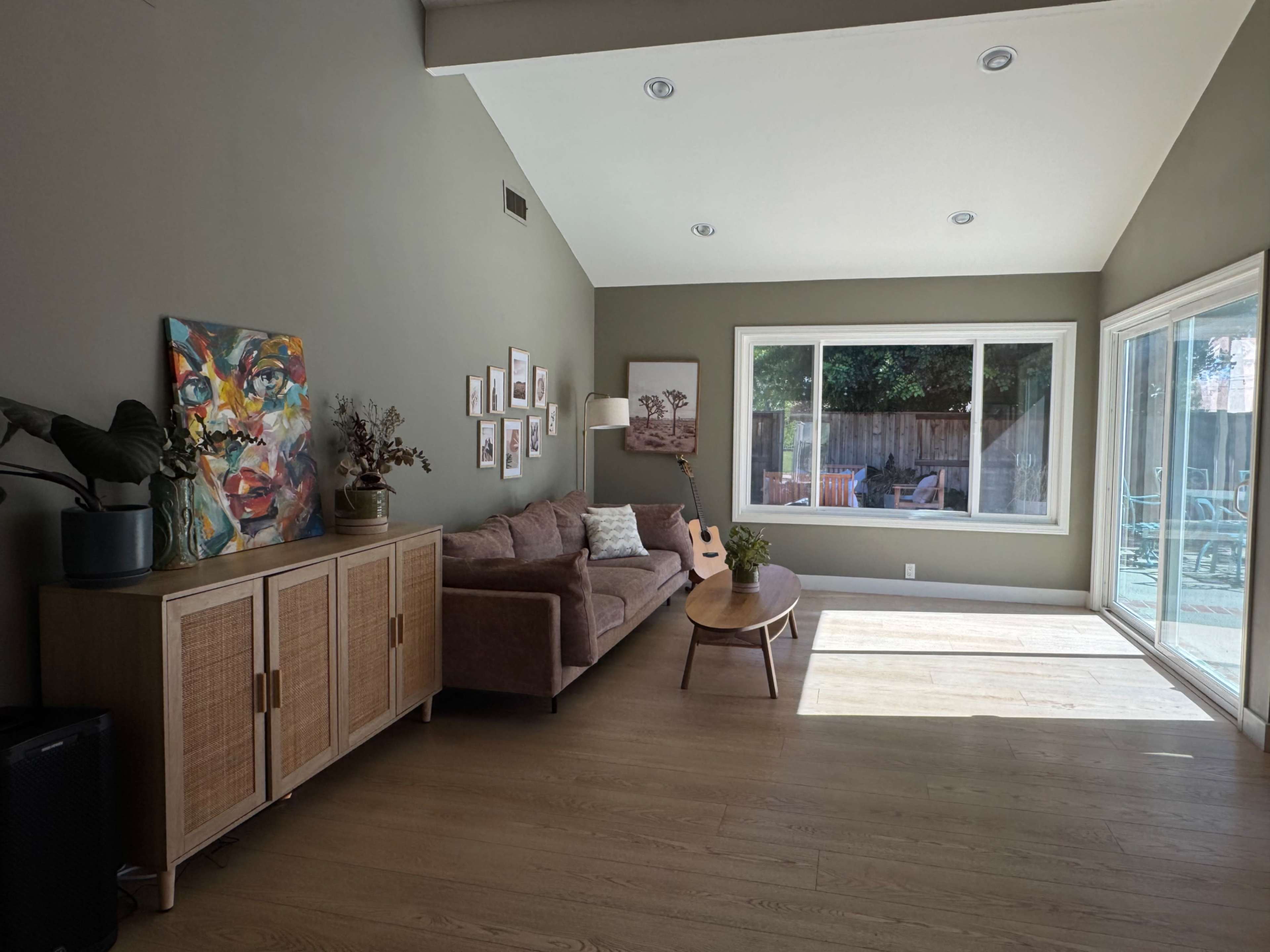 A modern living room features a sofa, wooden cabinet, wall art, and large windows allowing natural light.