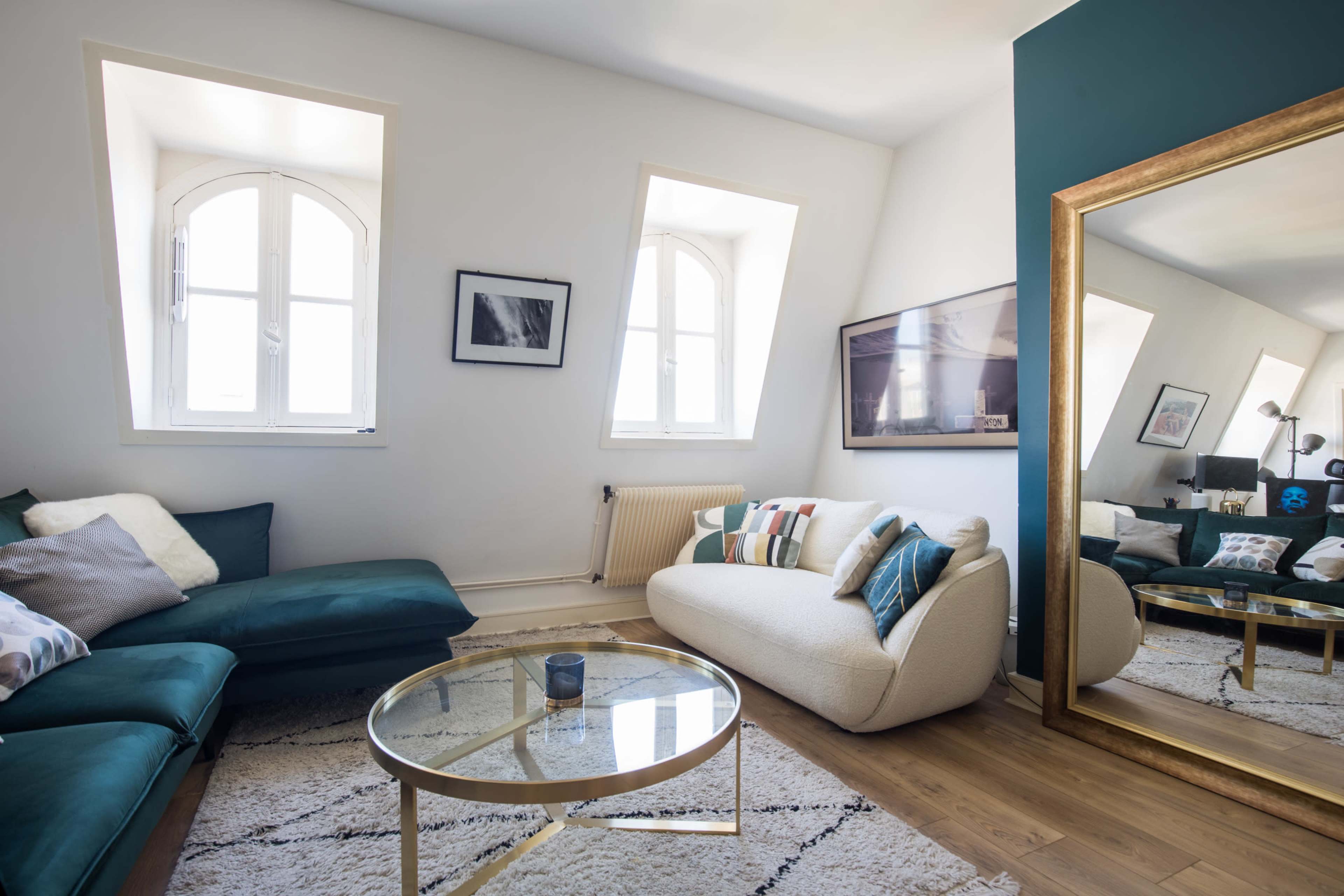 A modern living room features a plush sofa and armchair, a glass coffee table, and large windows with a mirror reflecting the space.
