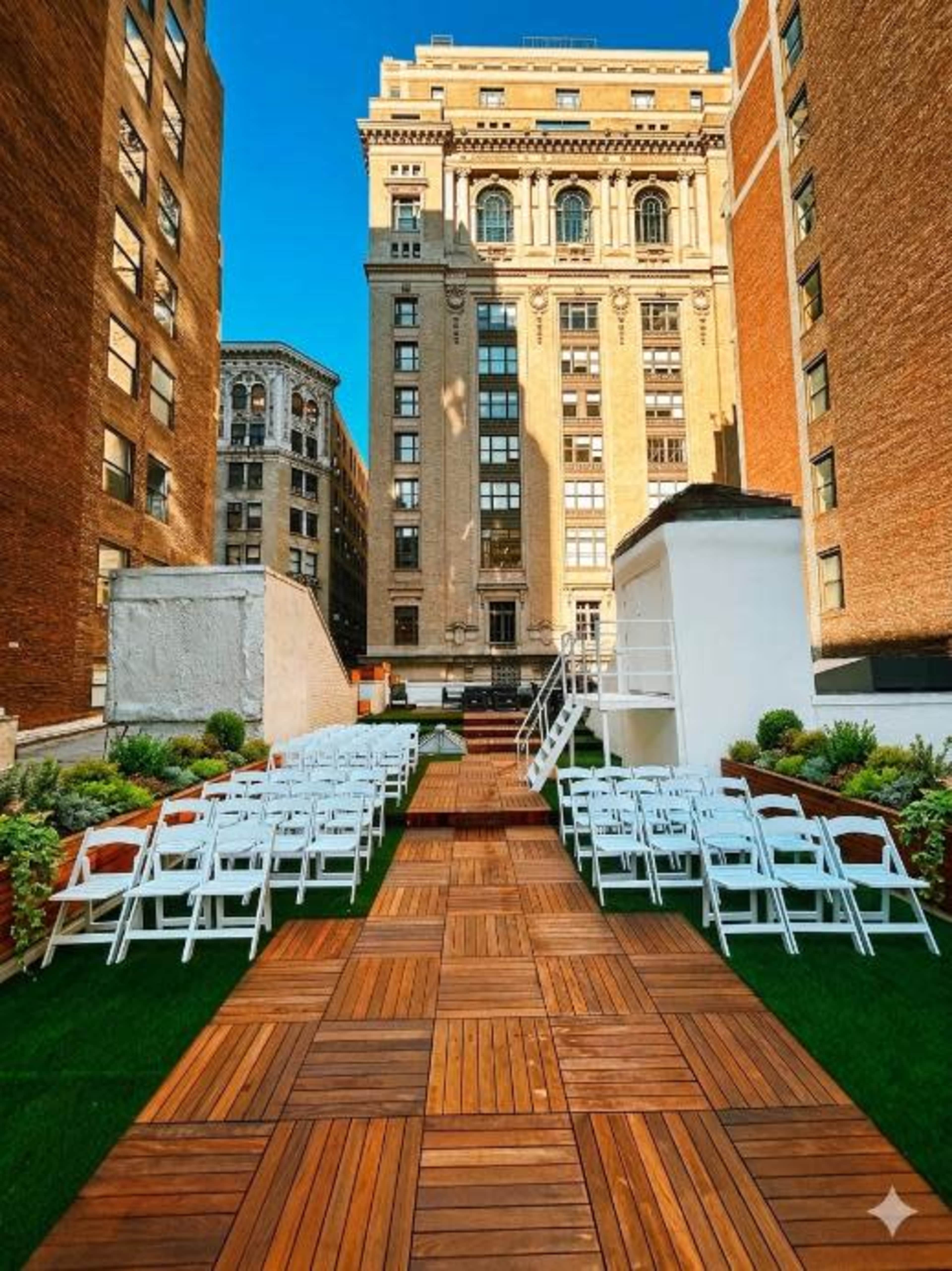 The image shows a rooftop event space with white chairs arranged in rows on a wooden walkway surrounded by greenery and tall buildings in the background.