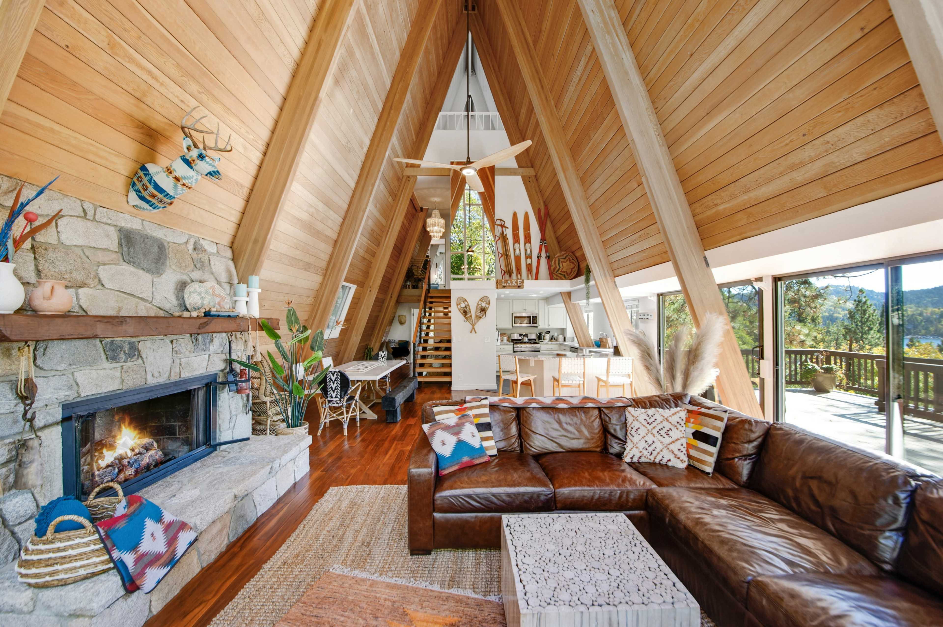 A modern A-frame cabin interior with a wooden framework, a stone fireplace, and a spacious living area featuring a leather sectional sofa and a view of the outdoors.
