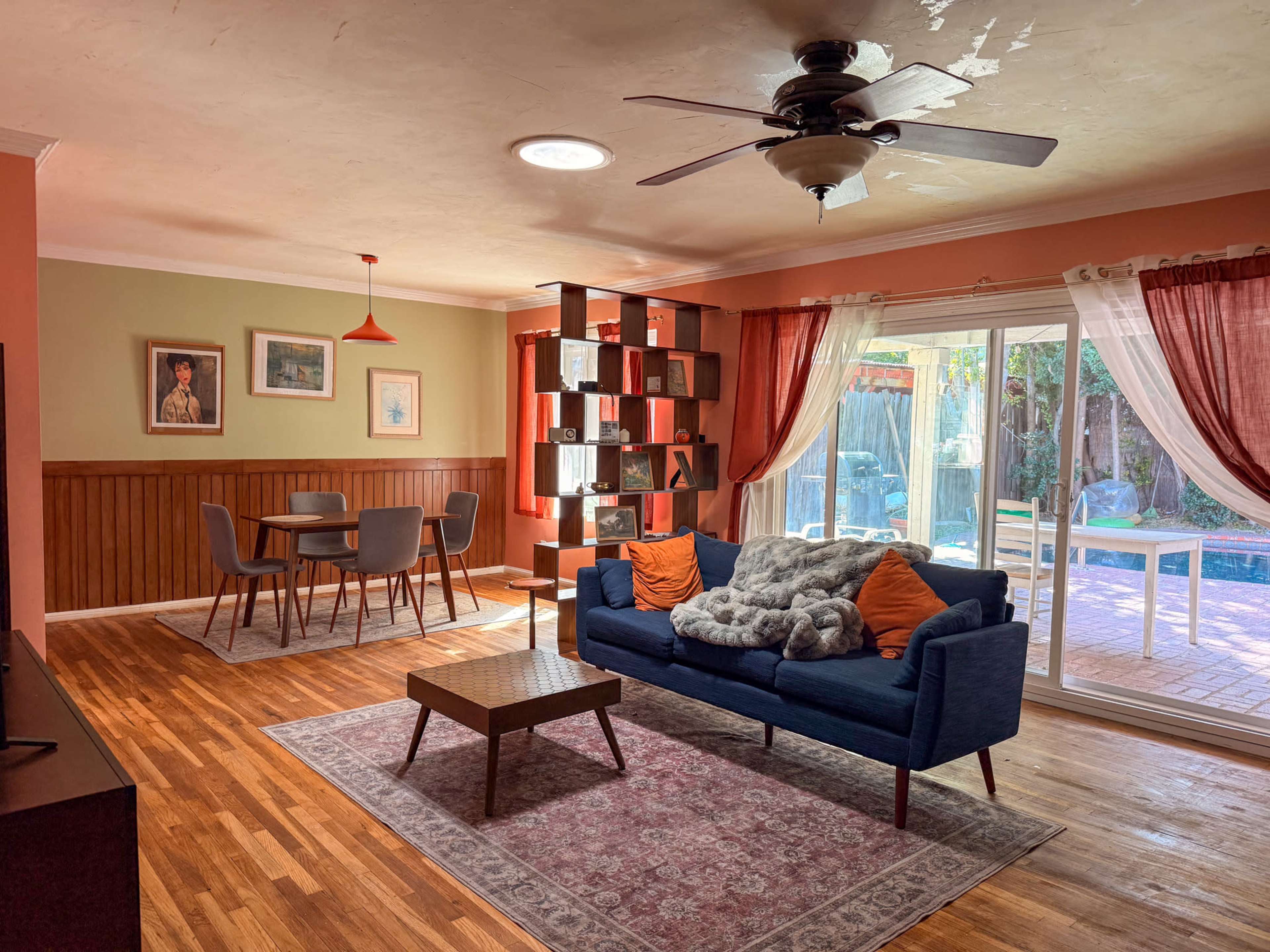 The living room features a blue sofa with orange cushions, a wooden coffee table, and a dining area with a round table and gray chairs, all illuminated by natural light from sliding glass doors.