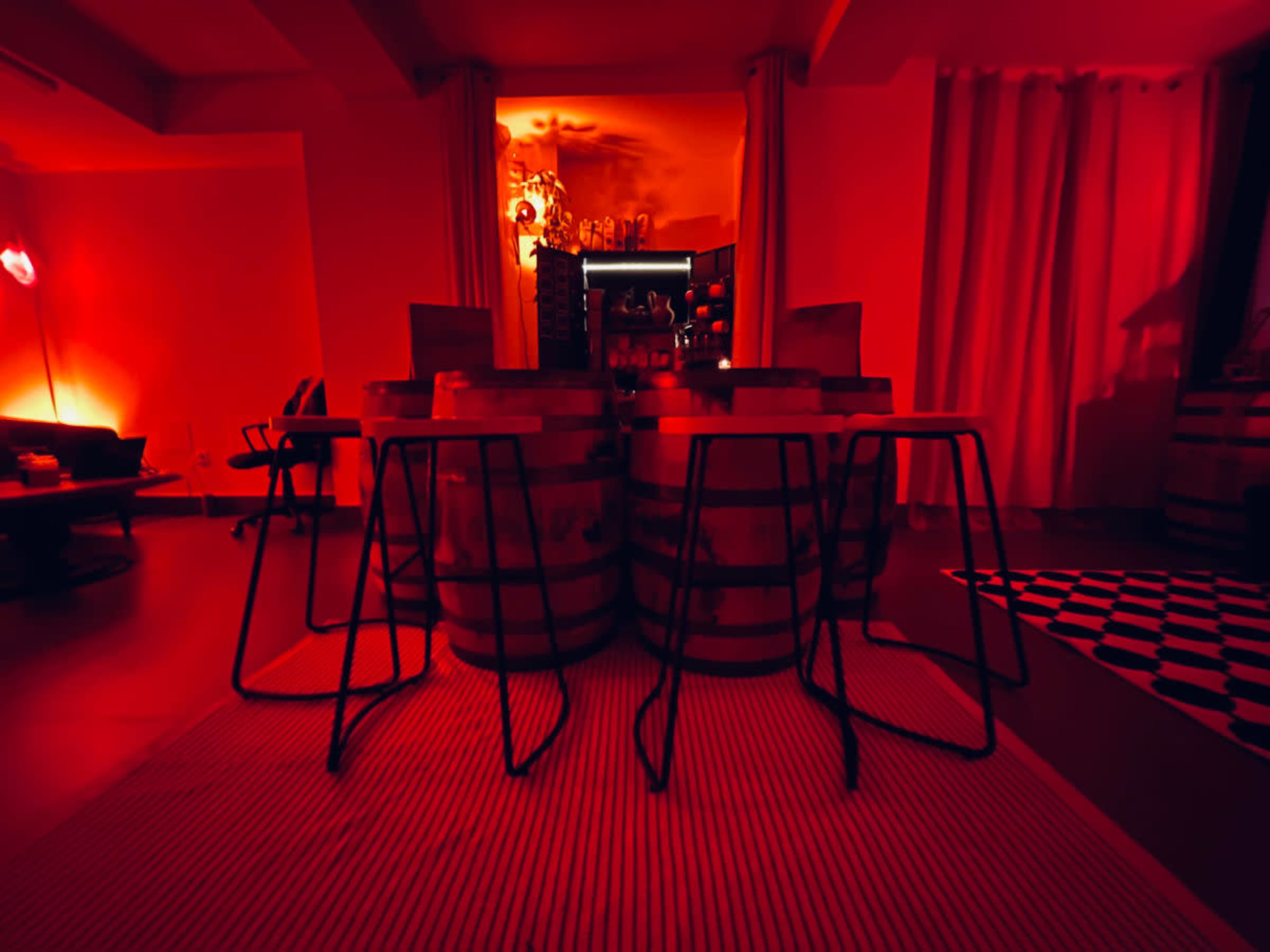 The image shows a room with red lighting featuring a bar made of wooden barrels and metal stools arranged around it.