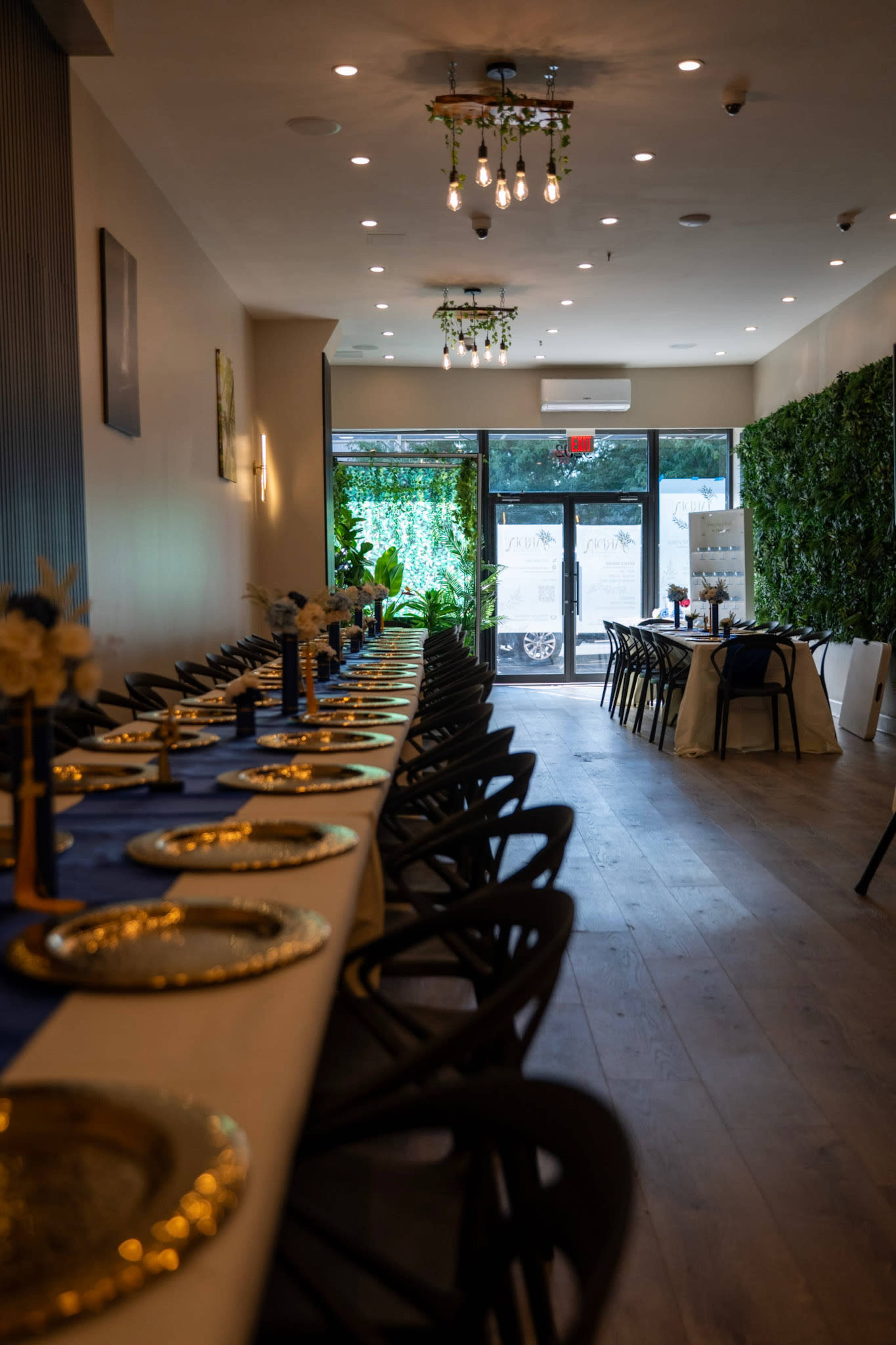 The image shows a restaurant interior featuring long tables set with elegant tableware, chandeliers overhead, and greenery lining the walls.