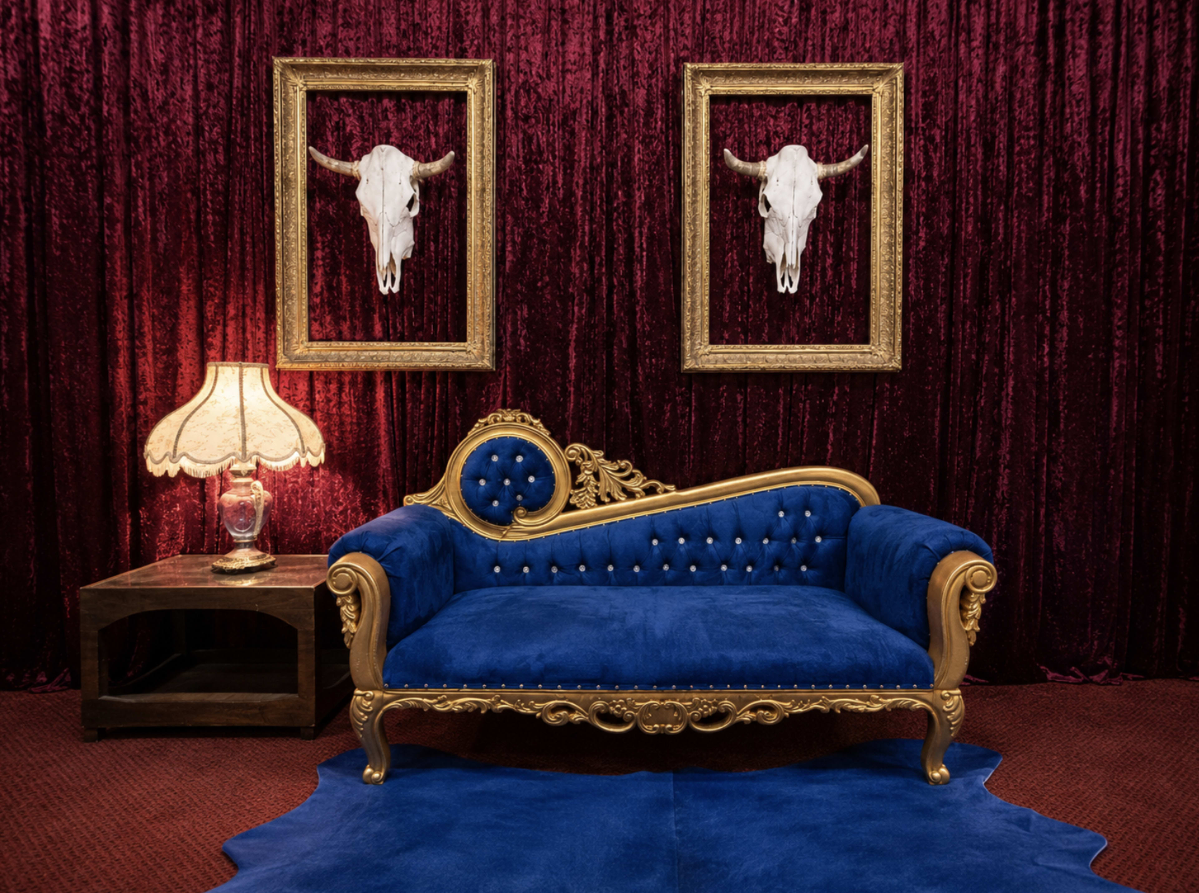 The image features a blue velvet sofa with ornate gold detailing, placed in front of a deep red velvet curtain, flanked by two framed cow skulls on the wall.