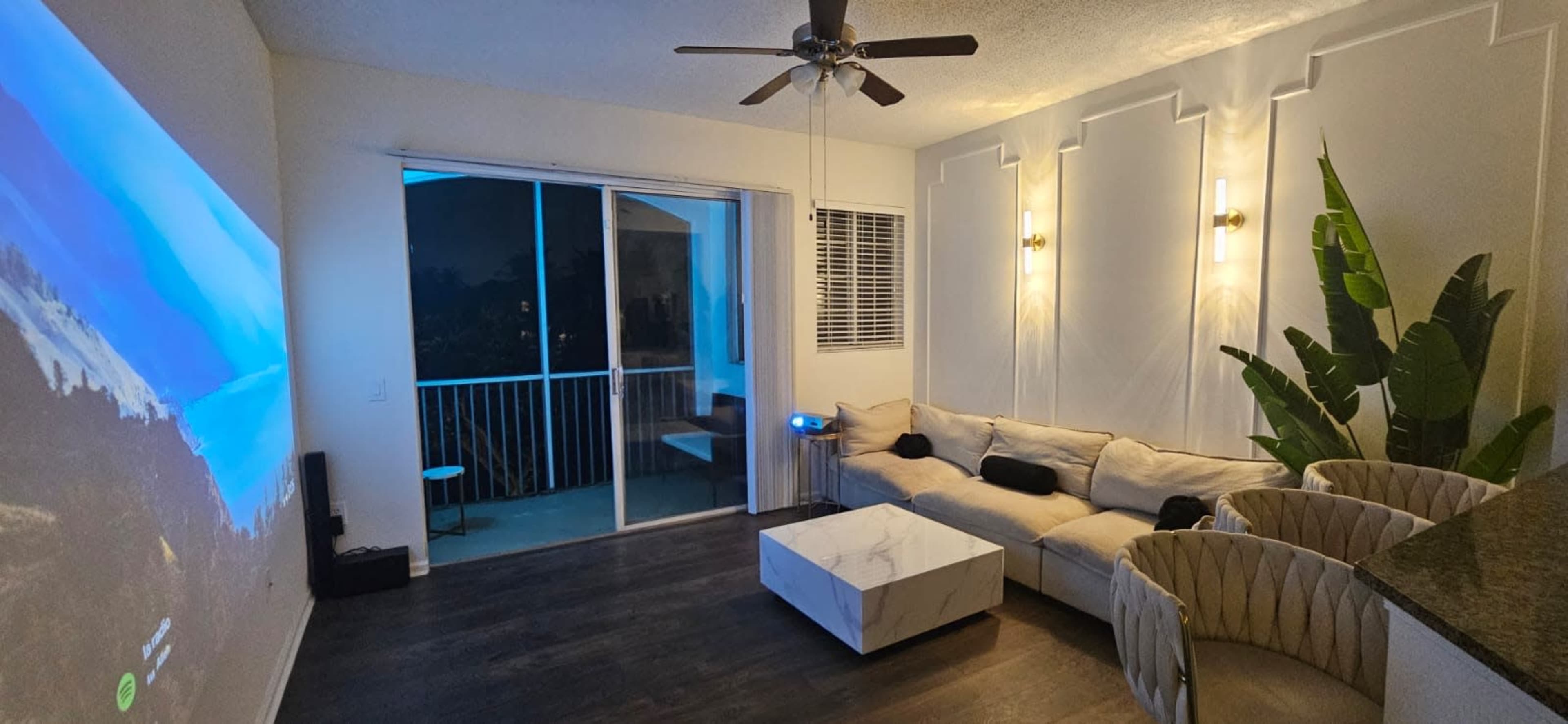 The image shows a modern living room with a large sectional sofa, a marble coffee table, and a projected image on the wall, alongside a balcony and potted plant.