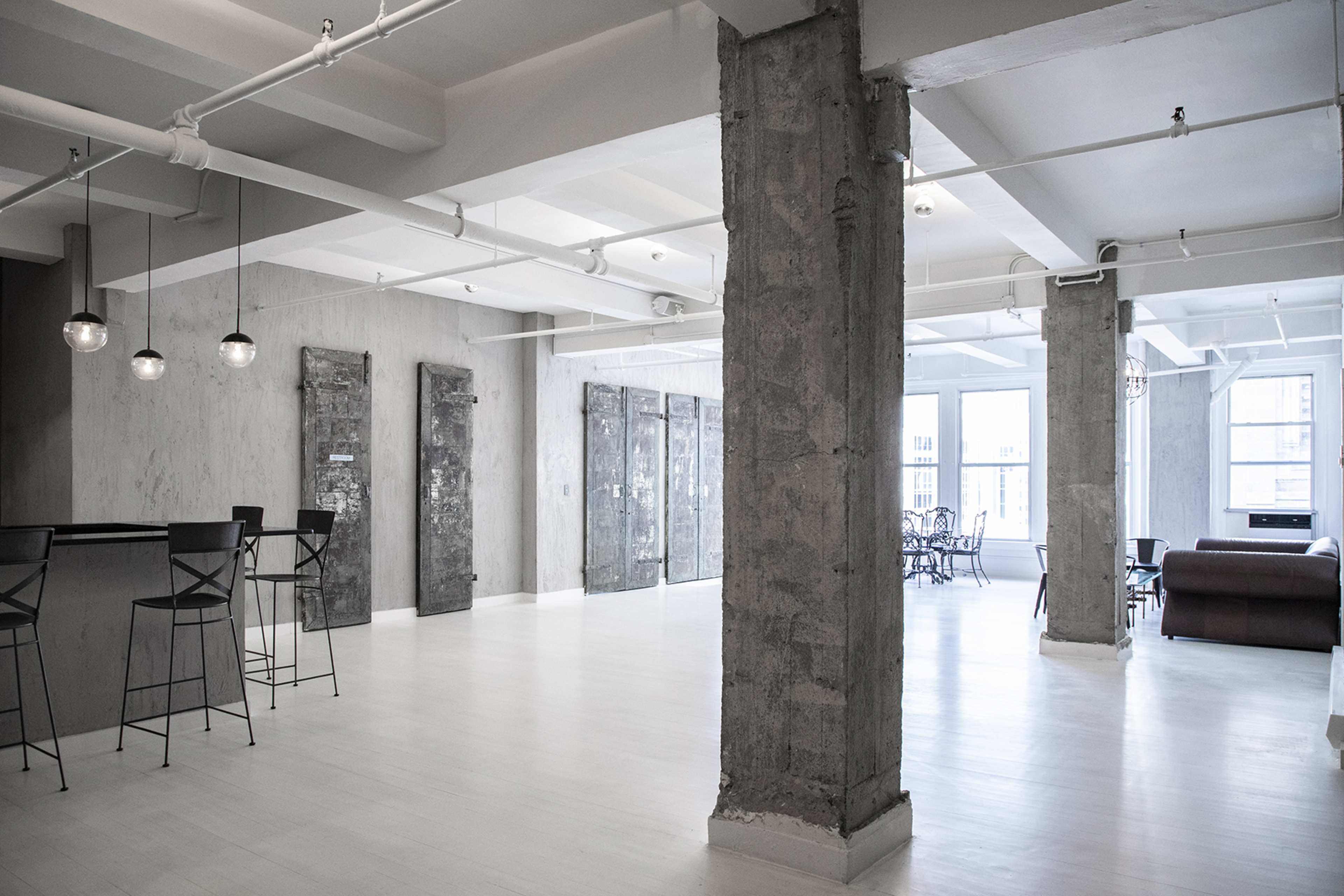 A spacious, industrial-style room featuring concrete pillars, a bar area, and a dining setup with chairs and a sofa.