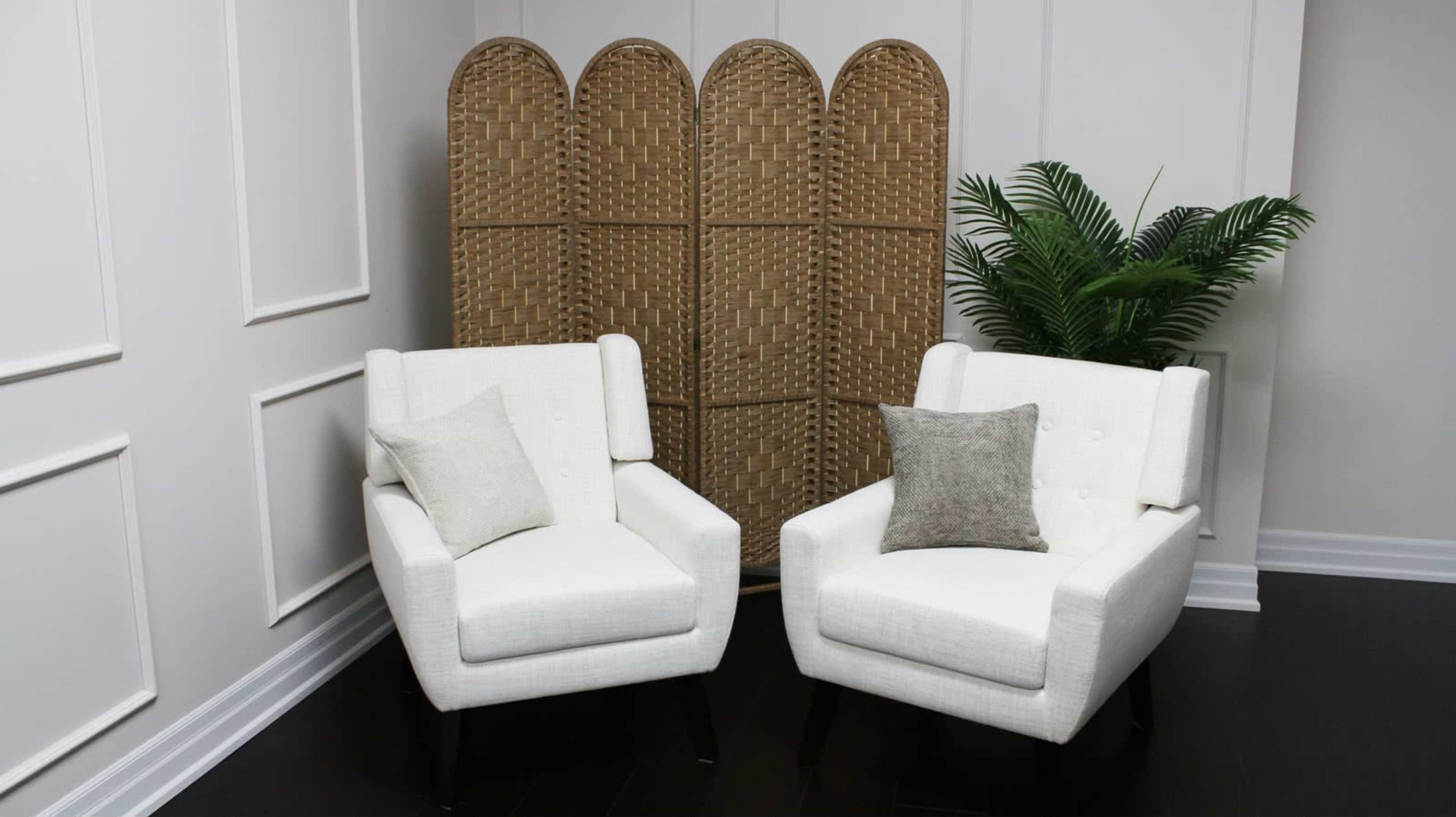 The image shows two white upholstered chairs with cushions, positioned opposite each other in front of a woven room divider and a potted plant.