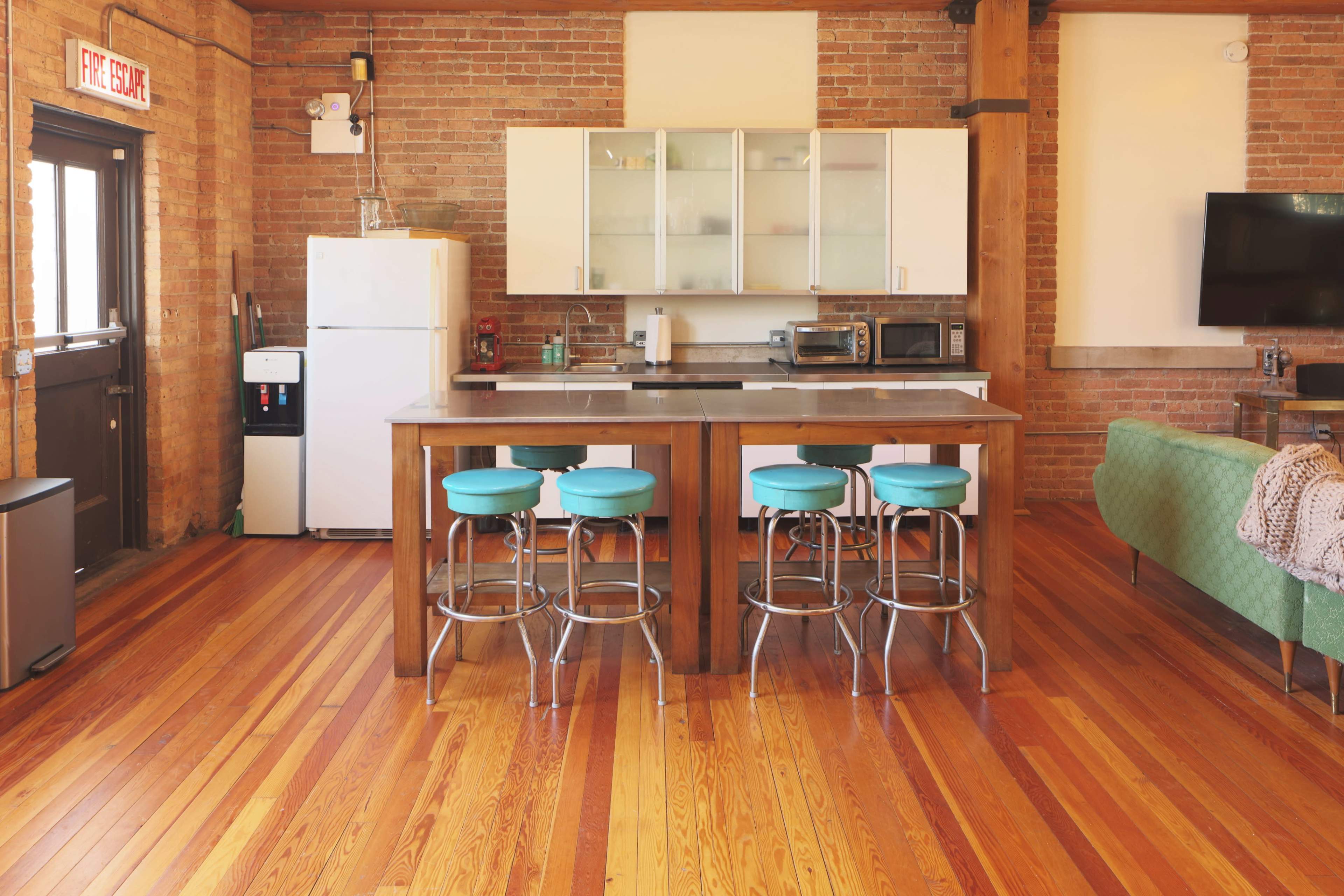The image shows a modern kitchen with a wooden island, turquoise bar stools, and stainless steel appliances, set against exposed brick walls and hardwood floors.