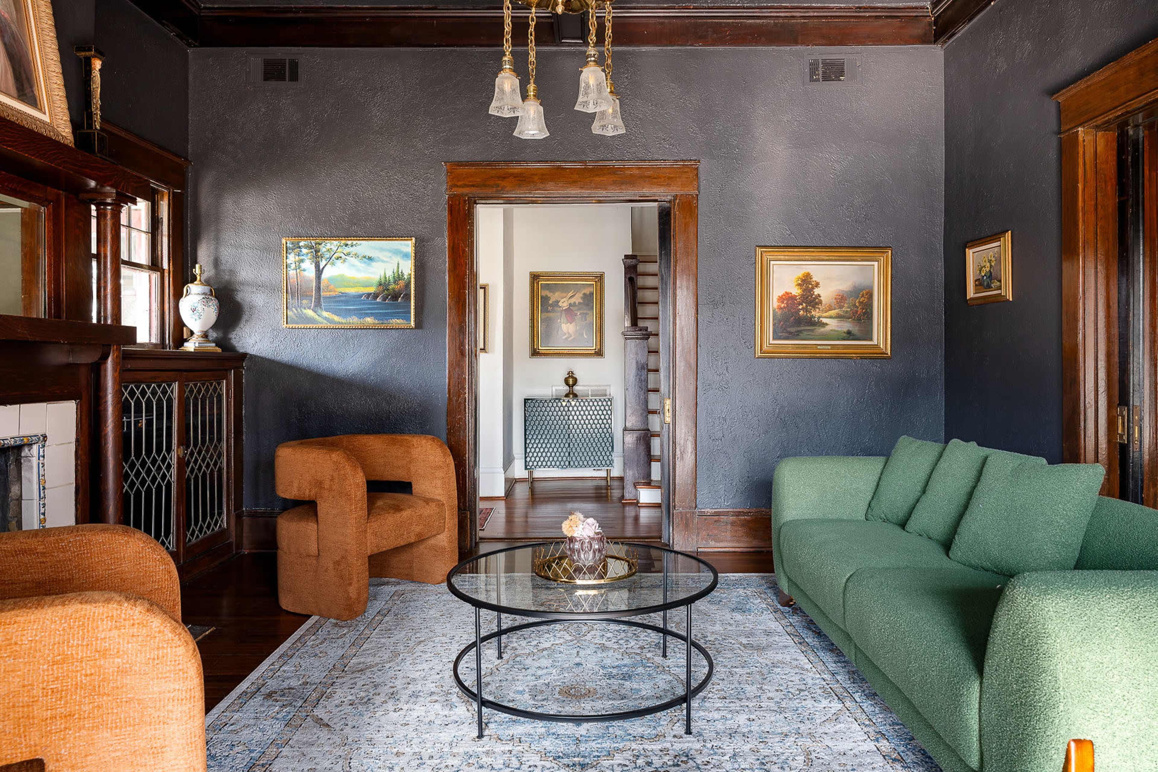 The image shows a stylish living room featuring a green sofa, two brown armchairs, a round glass coffee table, and framed paintings on the walls.