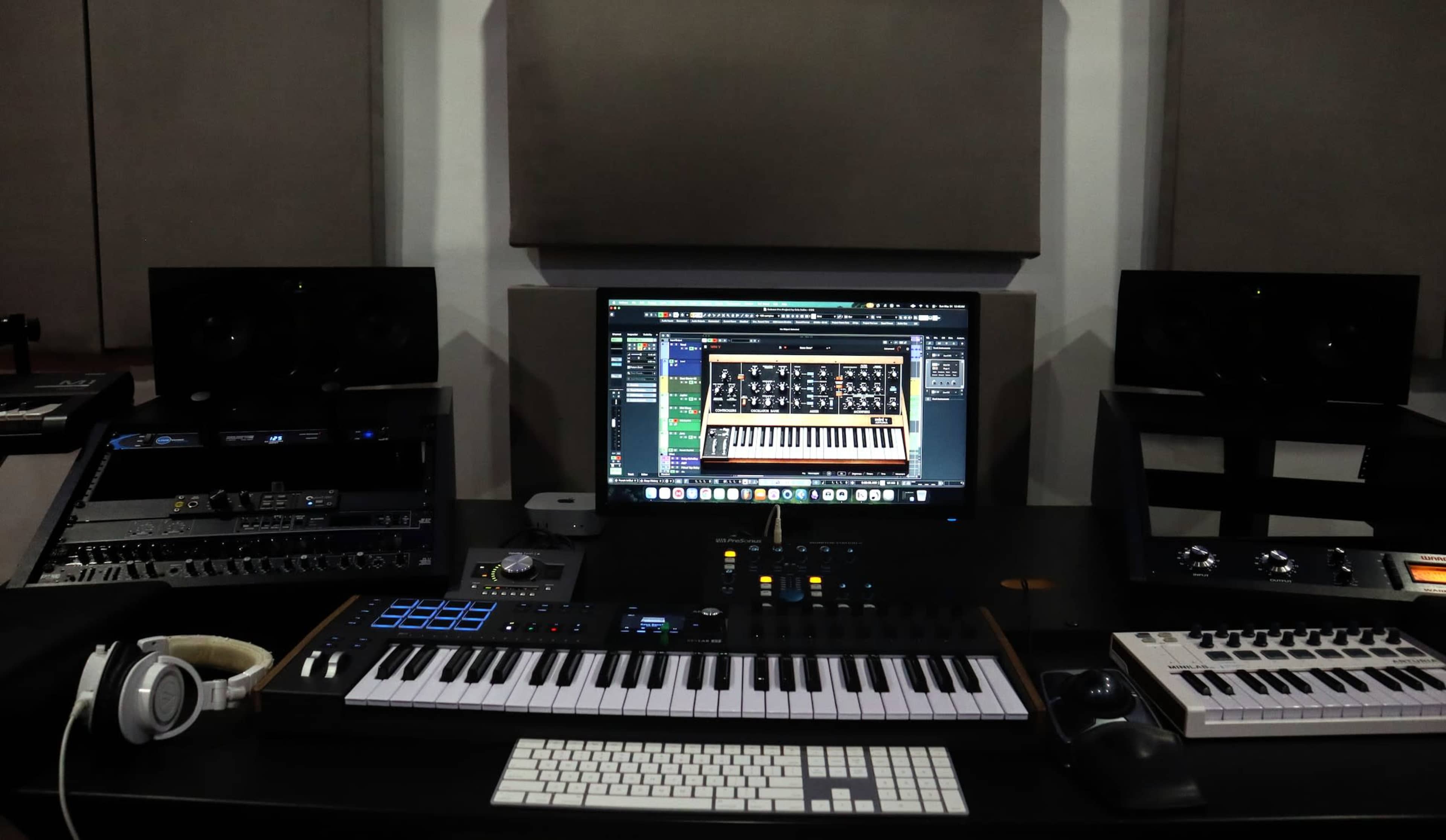 The image shows a music production setup with a computer monitor displaying software, a MIDI keyboard, studio speakers on either side, and various audio equipment on a black desk.