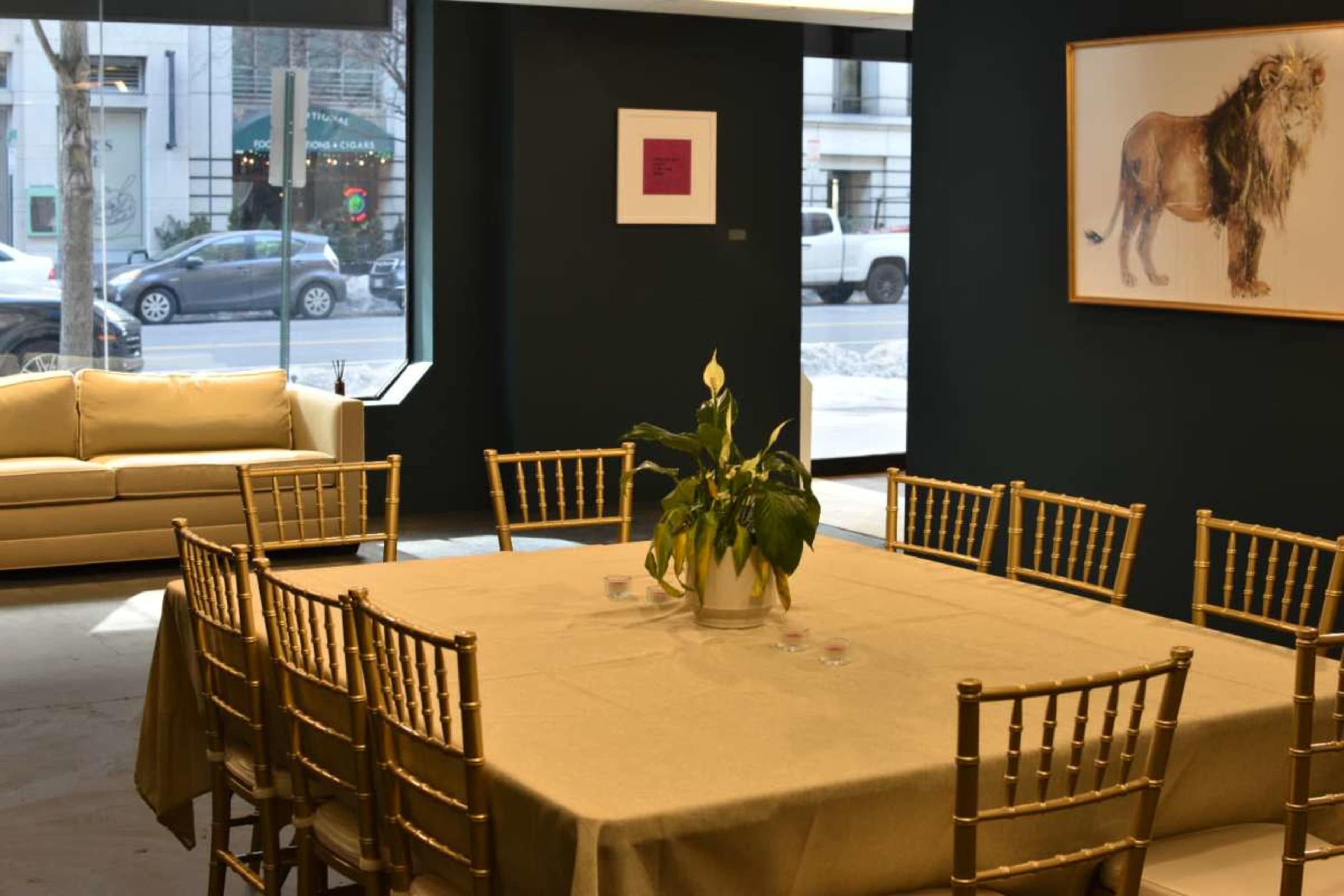 The image shows a modern interior space with a large table surrounded by gold chairs, a potted plant in the center, and a couch along the wall, with a lion artwork displayed nearby.
