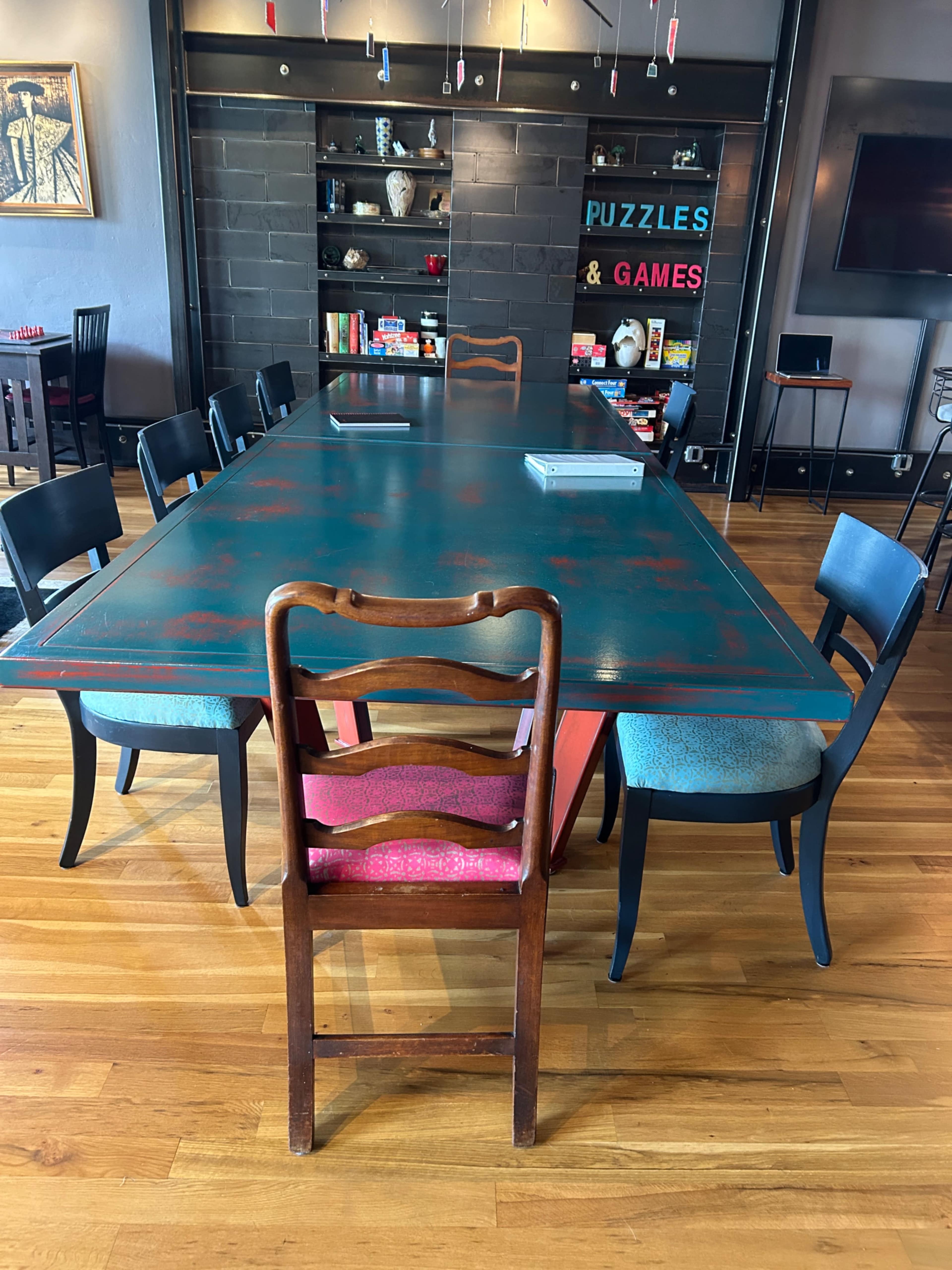 A long, teal table with a weathered finish is surrounded by various chairs, including a prominent wooden one at the head, in a room filled with shelves displaying games and decorative items.