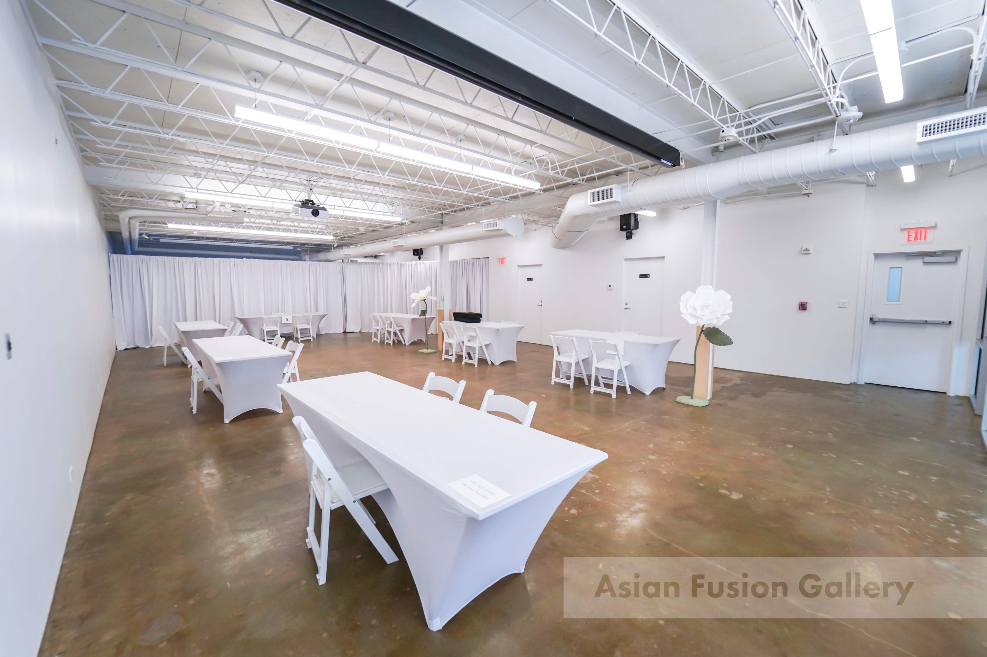 A spacious, minimalist event hall with white walls, several rectangular tables covered in white tablecloths, and a large decorative flower display at one end.