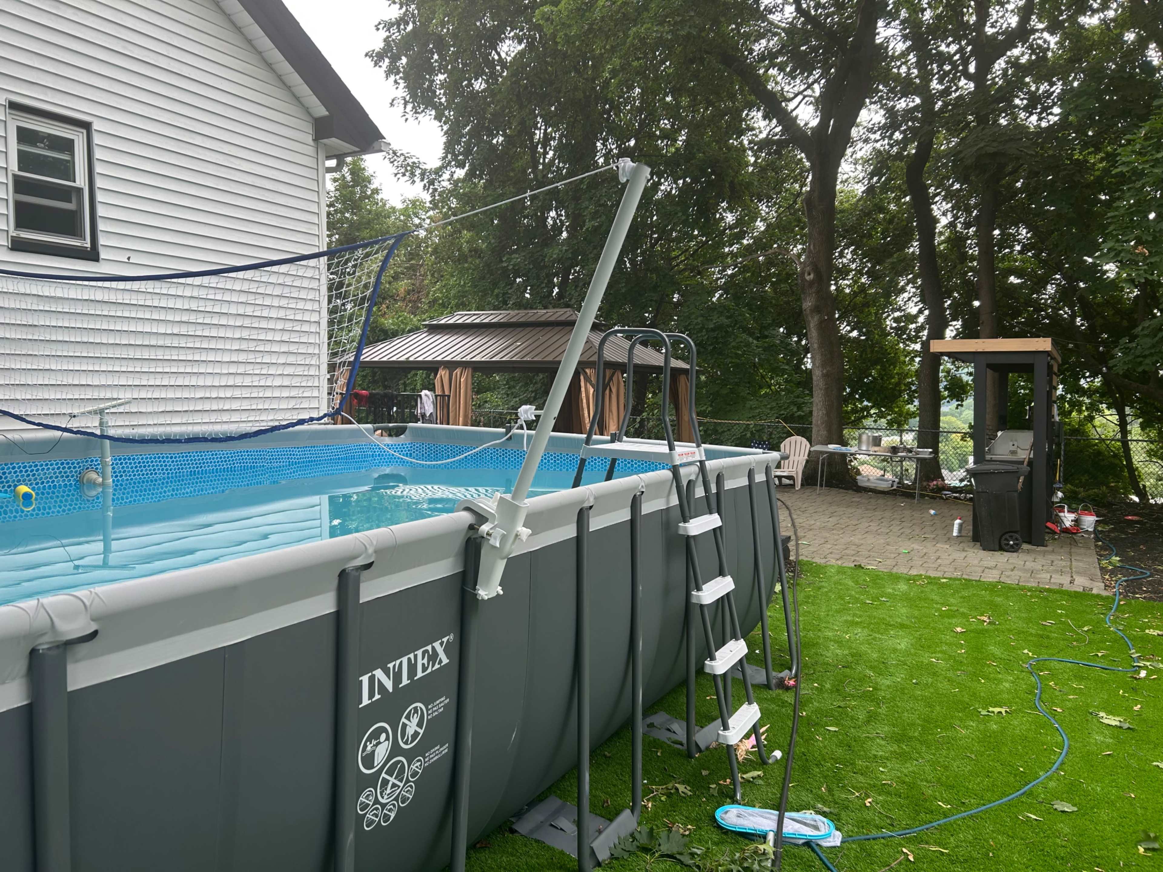 The image shows a rectangular above-ground swimming pool set in a backyard with a grassy area, surrounded by trees and a patio with a gazebo.