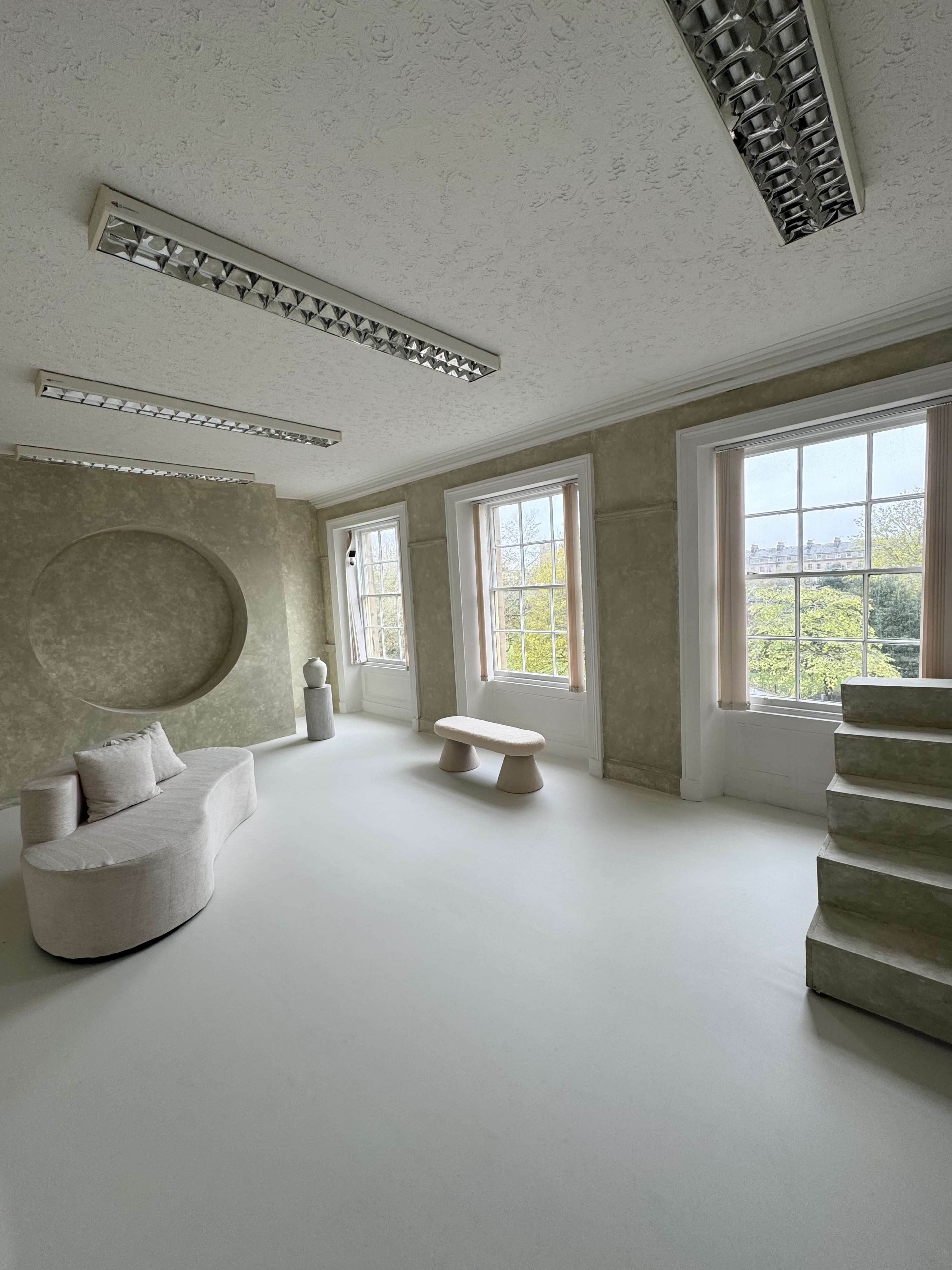 The image shows a spacious, minimalist room with large windows, a light-colored curved sofa, a sculptural table, and a staircase leading to another level.