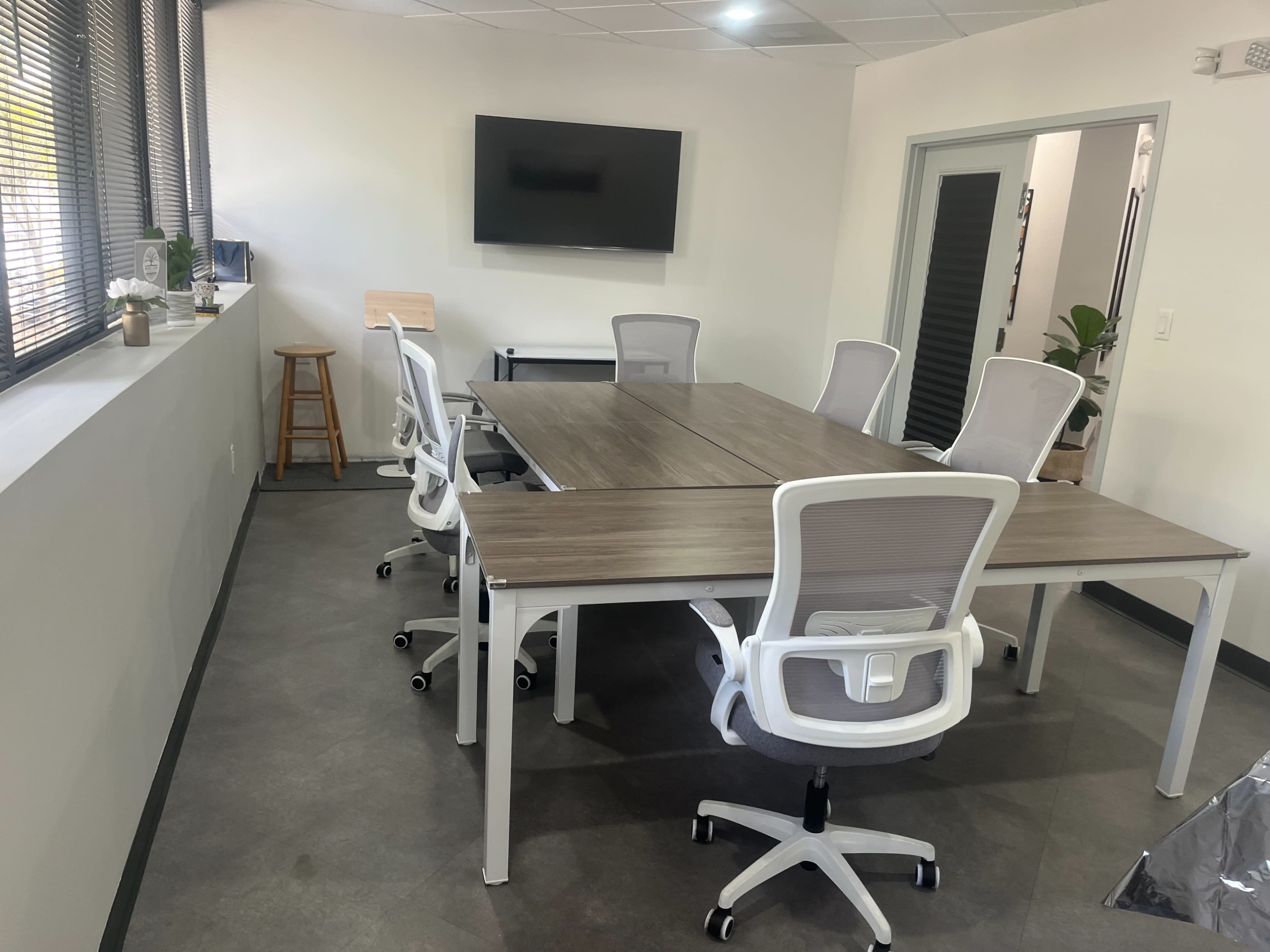 The image shows a modern conference room with a large wooden table surrounded by six white rolling chairs, a television on the wall, and a window with blinds.