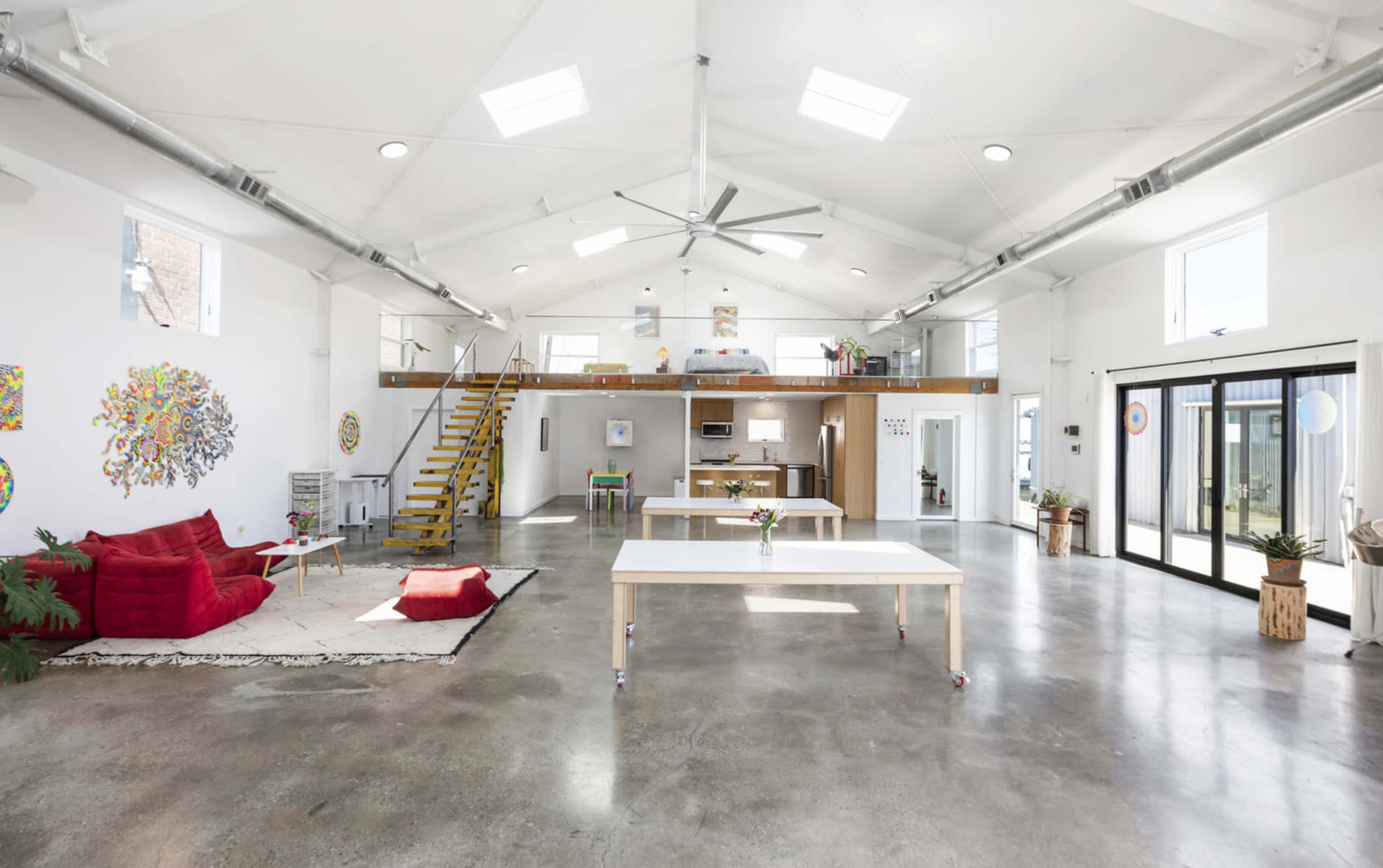 The image shows a spacious, open-concept interior with high ceilings, featuring a living area with red seating, a kitchen area on a raised platform, and large glass doors leading to an outdoor space.