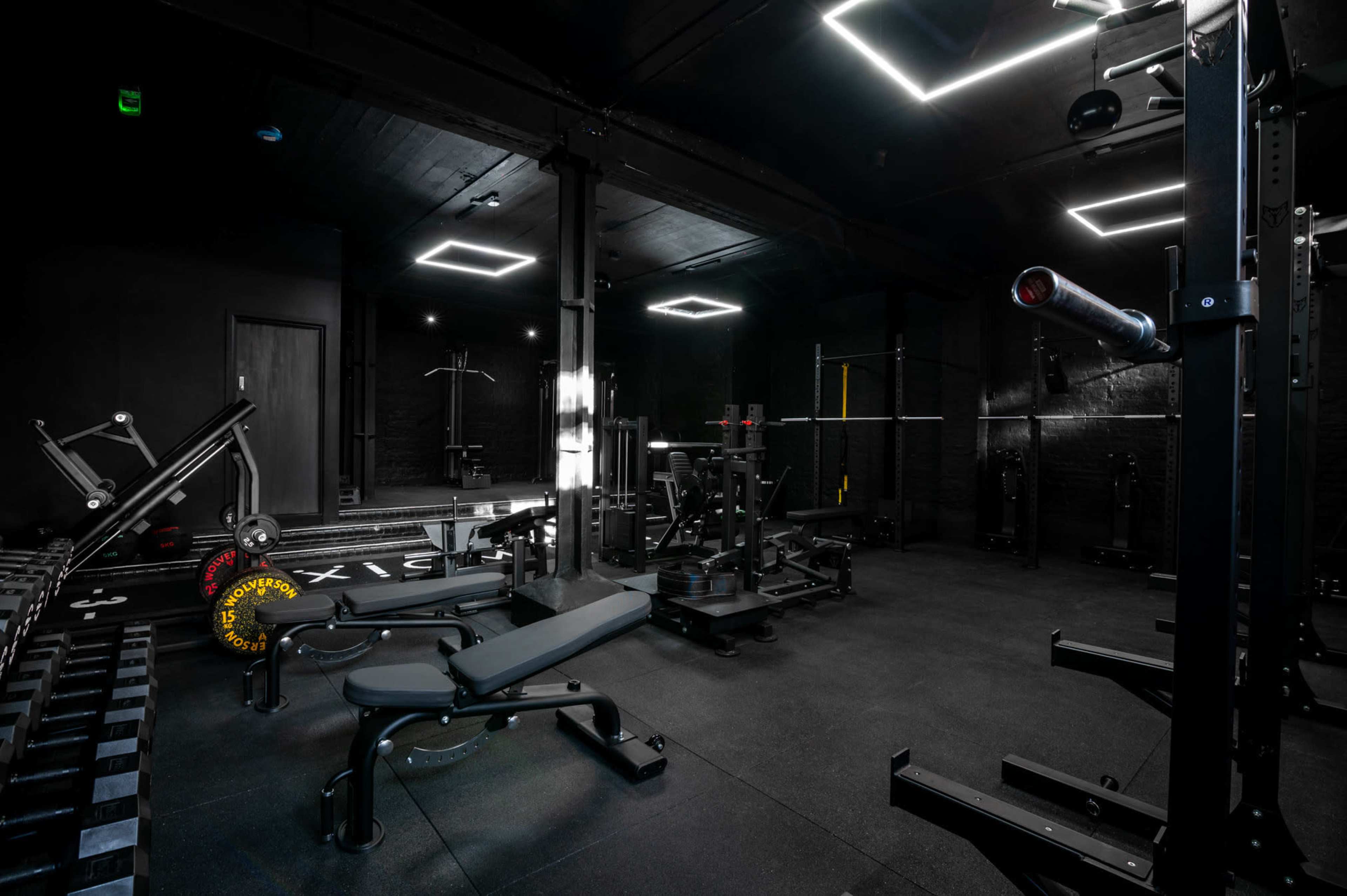 The image shows a dimly lit gym with various weightlifting equipment and benches arranged on a black floor.