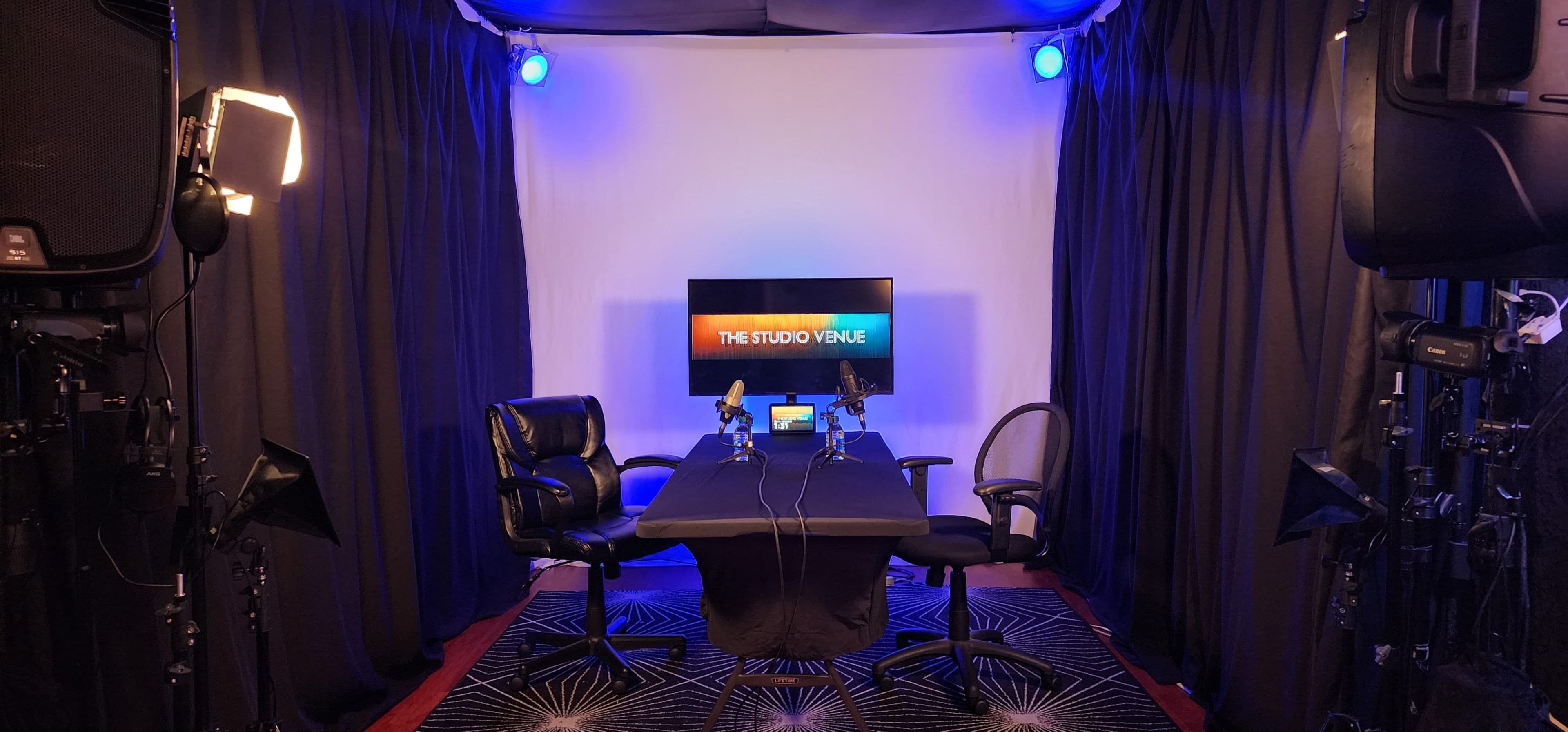 The image shows a recording studio setup with two chairs flanking a table, a backdrop of black curtains, and a screen displaying the text "THE STUDIO VENUE."