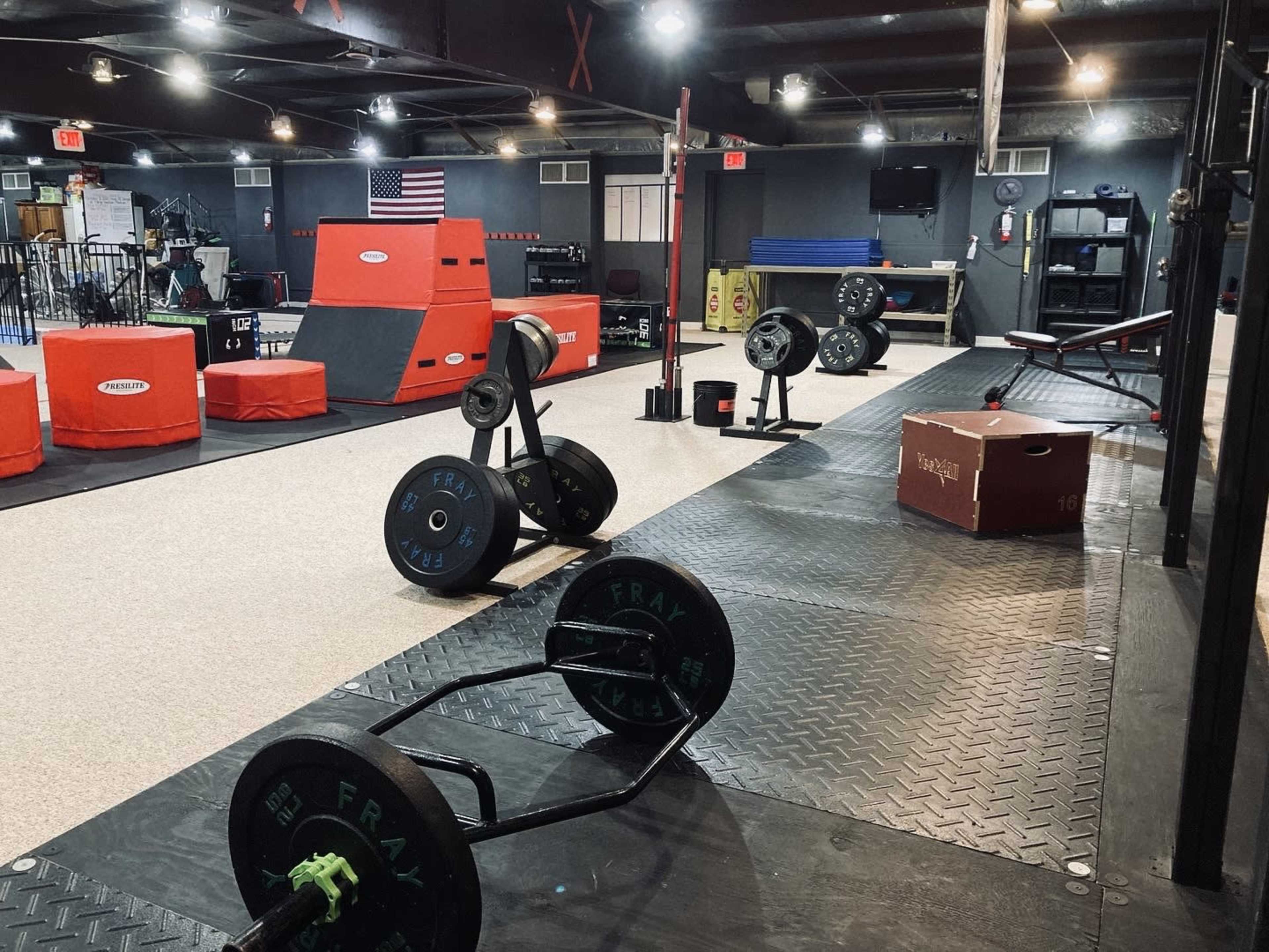 A gym interior features various weightlifting equipment, including barbell racks and exercise mats, alongside a functional training area with obstacles.