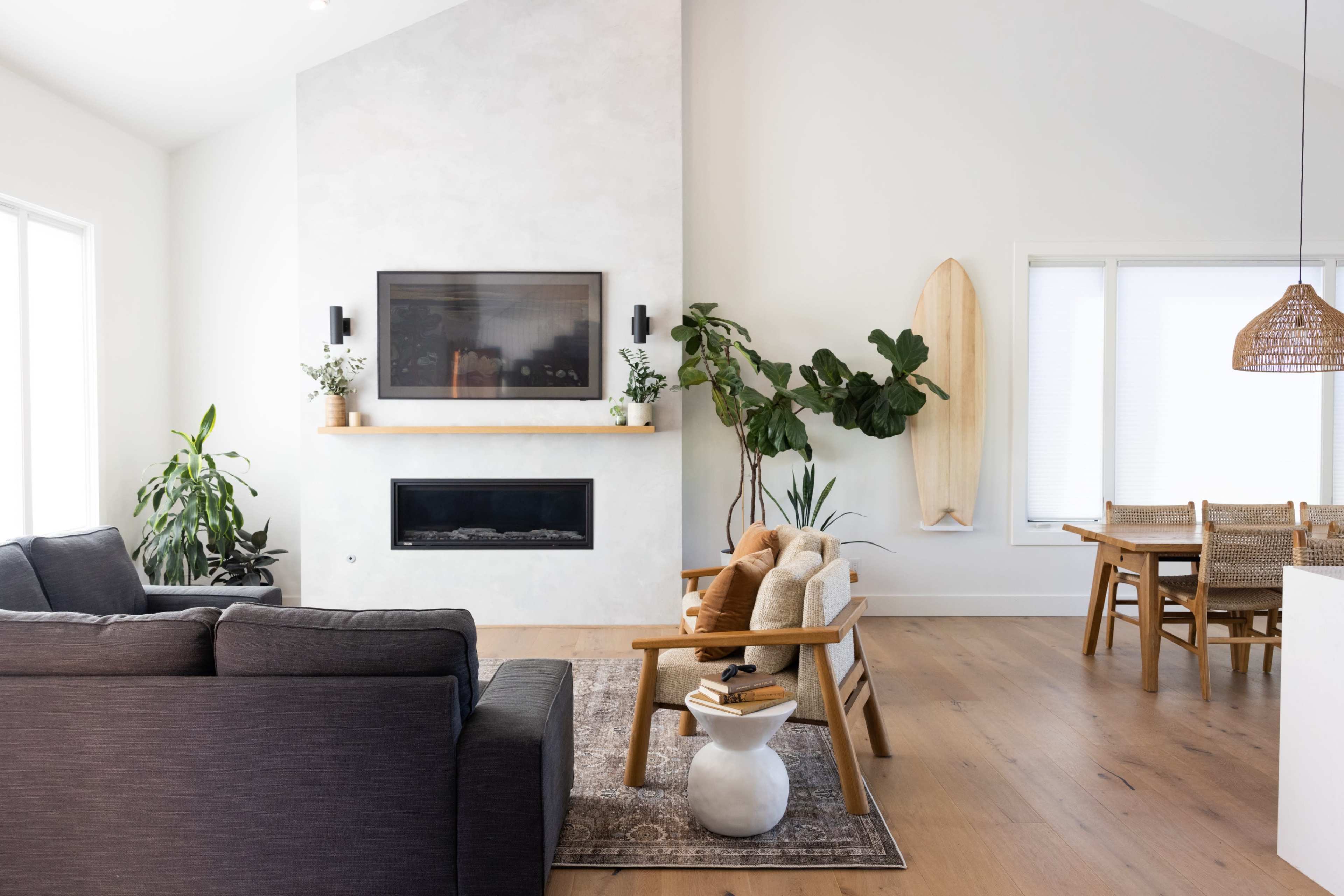 A modern living room featuring a sectional sofa, a coffee table with stacked books, and a dining area with wooden chairs, accompanied by plants and a surfboard on the wall.